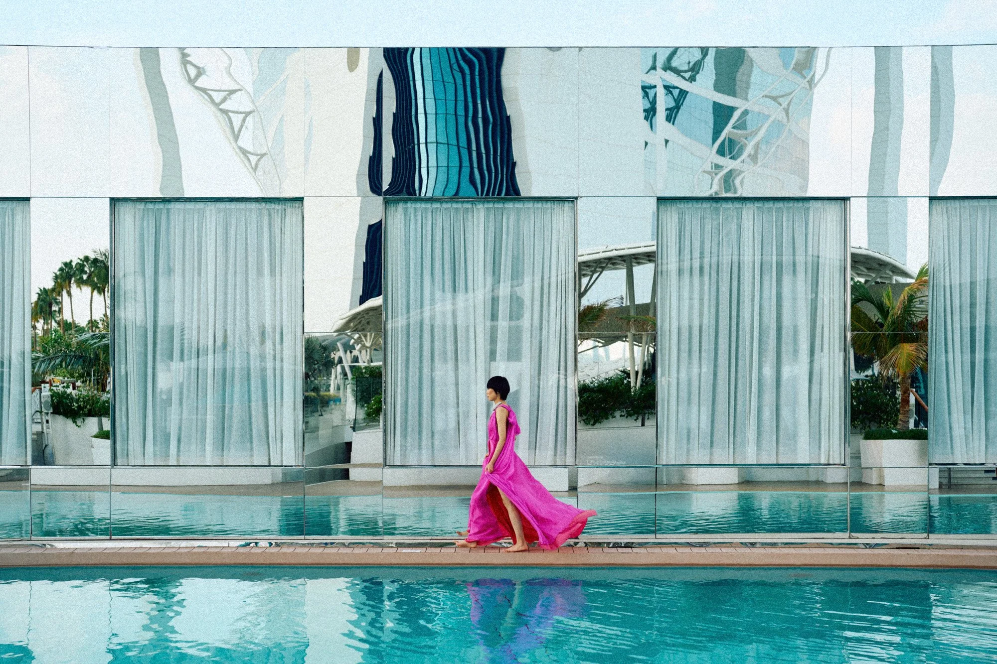 A woman in a bright pink dress walking on a poolside walkway reflects in the water, with a modern building featuring glass windows and curtains, and palm trees in the background.
