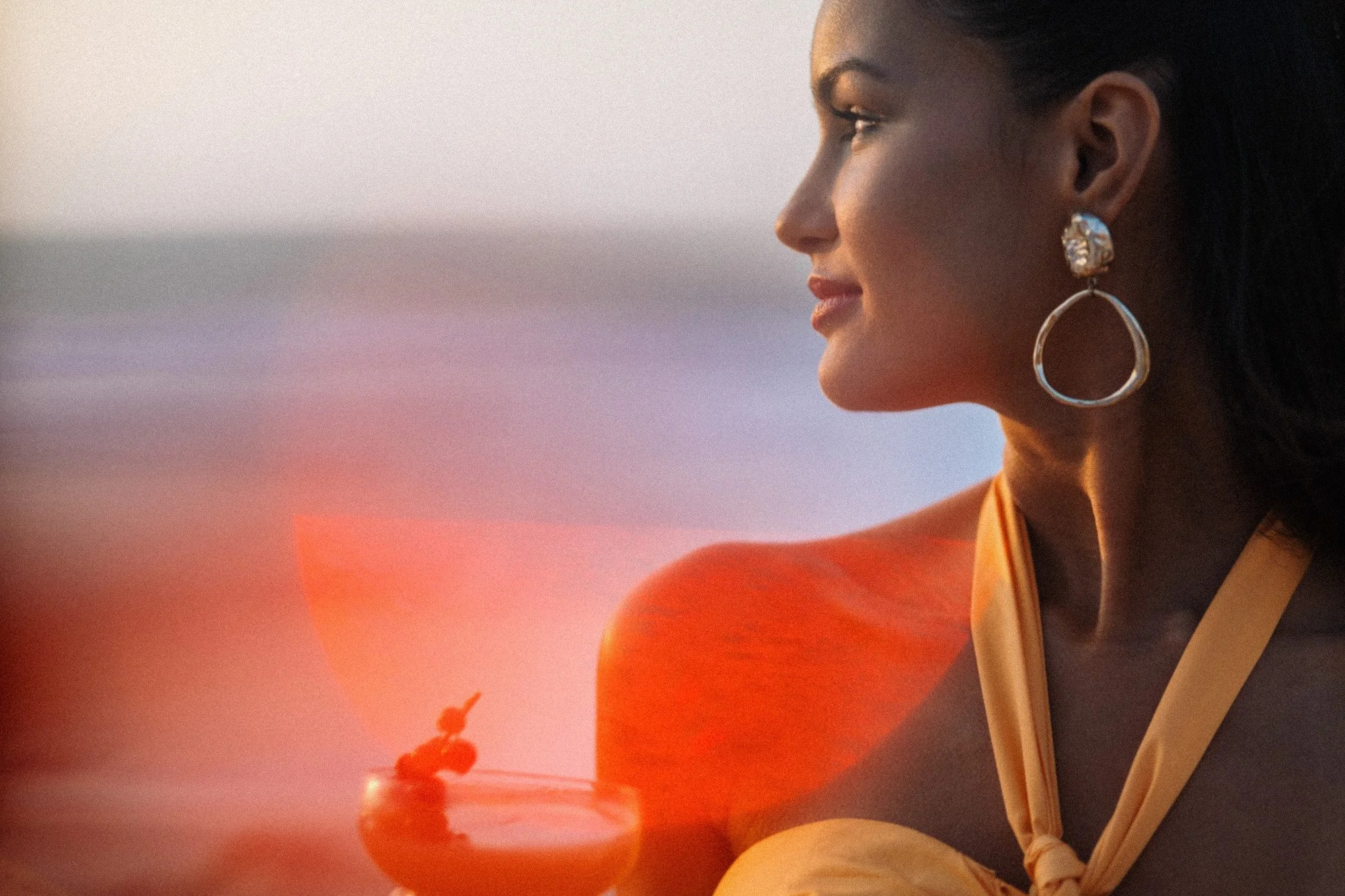 A woman with dark hair and earrings looking to the side with a sunset in the background.