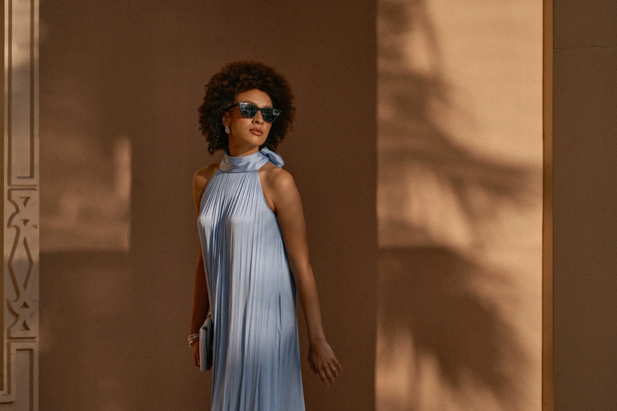A woman with curly hair wearing sunglasses and a light blue pleated dress with a high neck and scarf detail, holding a clutch, standing against a beige wall with shadow patterns.