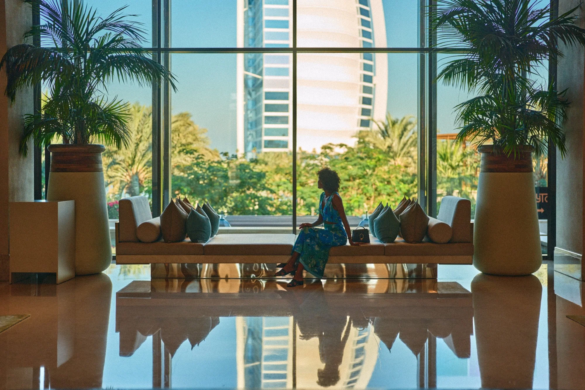 A woman sitting on a bench in a modern hotel lobby with large glass windows, palm trees inside planters, and a cityscape with a tall distinctive building outside.