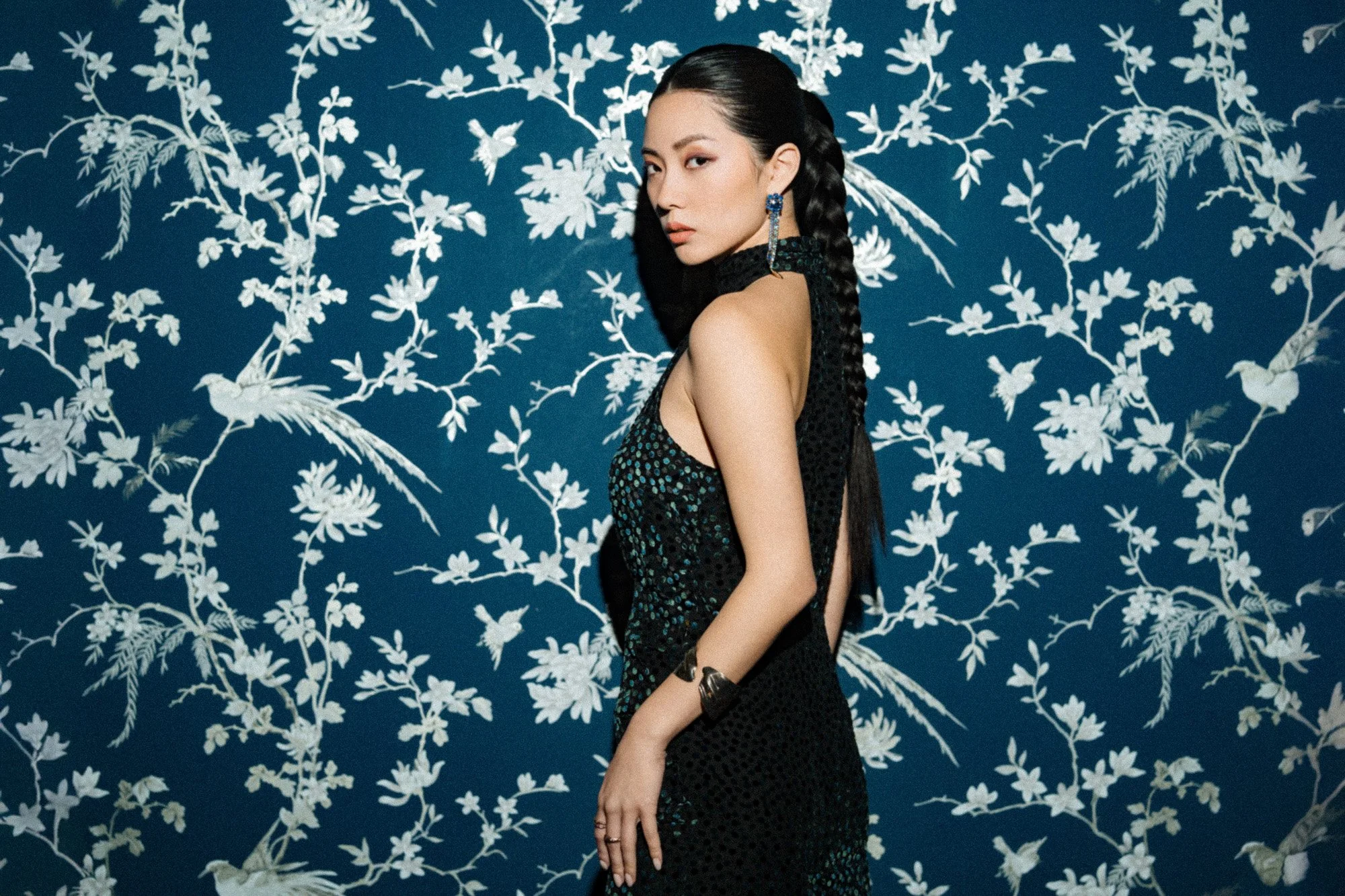 A woman with long black braided hair wearing a black sleeveless dress with a high slit, standing against a blue wall with a white floral and bird pattern.
