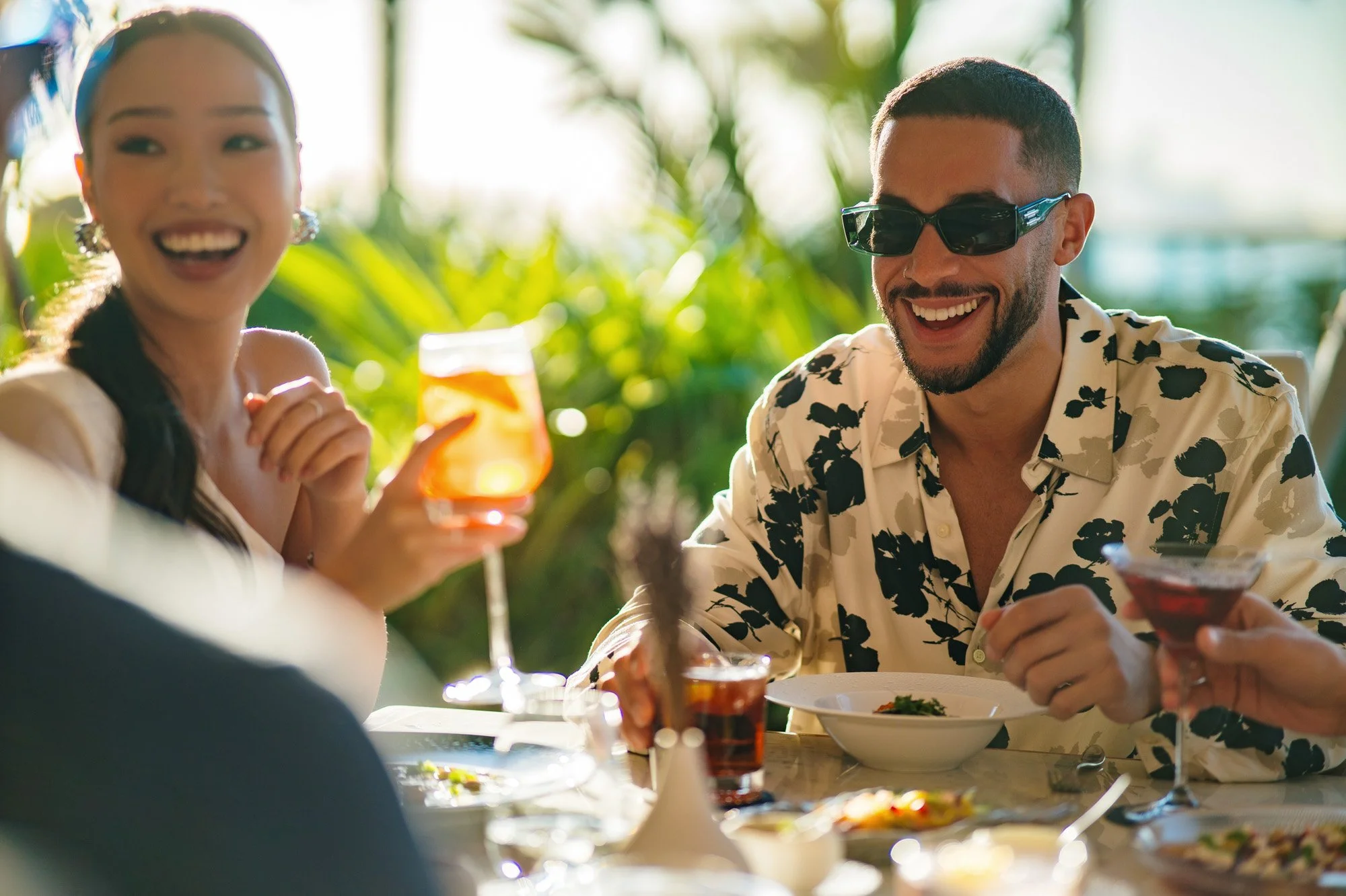 People enjoying drinks and food at an outdoor restaurant with greenery in the background.