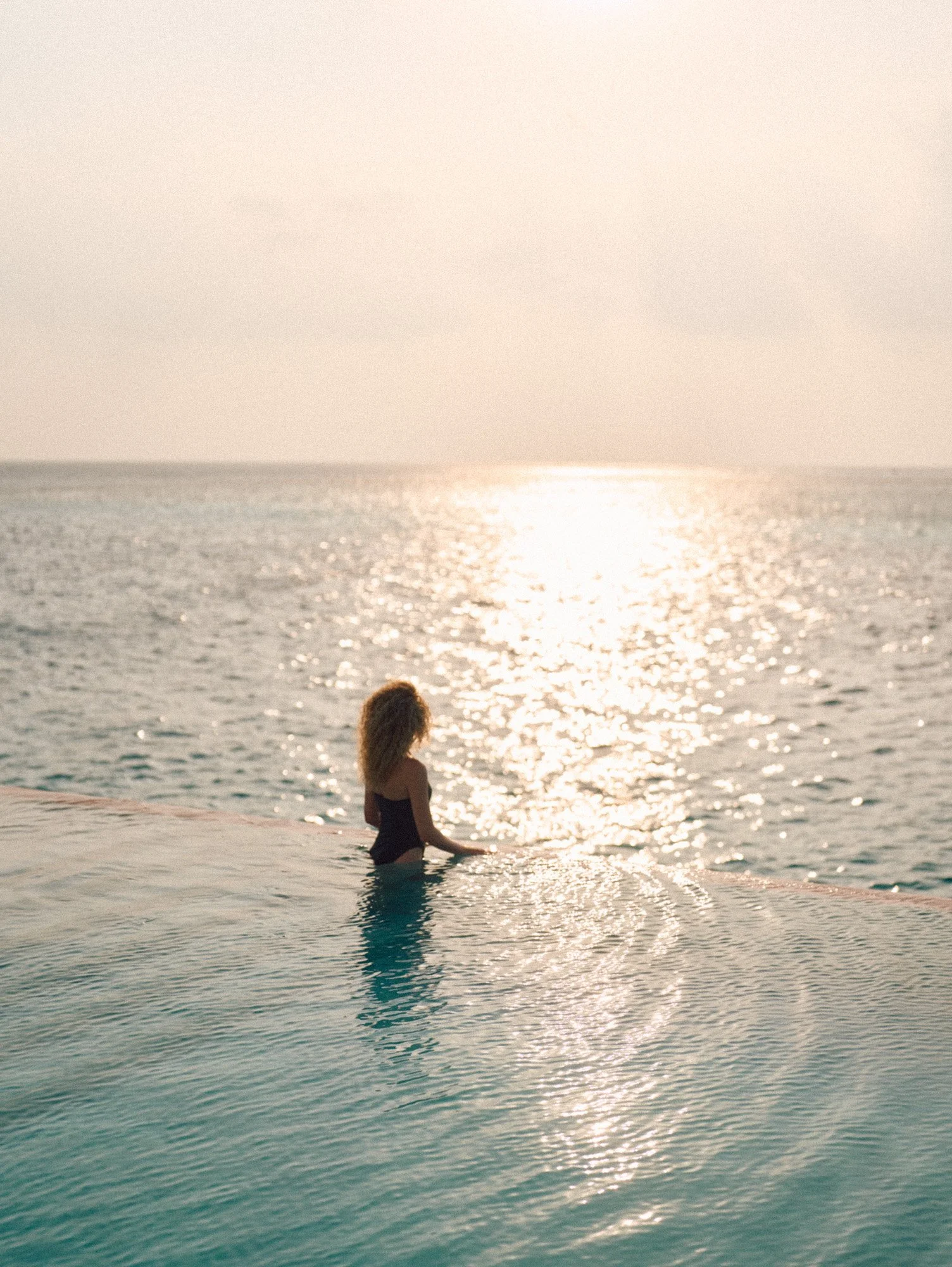 A woman with curly hair standing in an infinity pool overlooking the ocean during sunset.