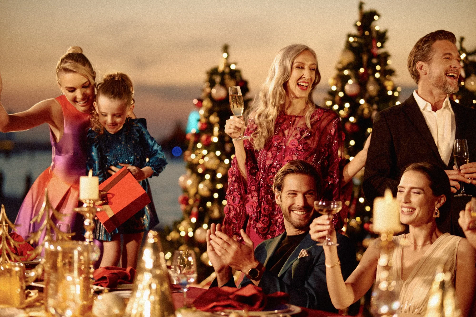 A group of people celebrating Christmas outdoors by a decorated Christmas tree, with some holding glasses of champagne, during sunset.