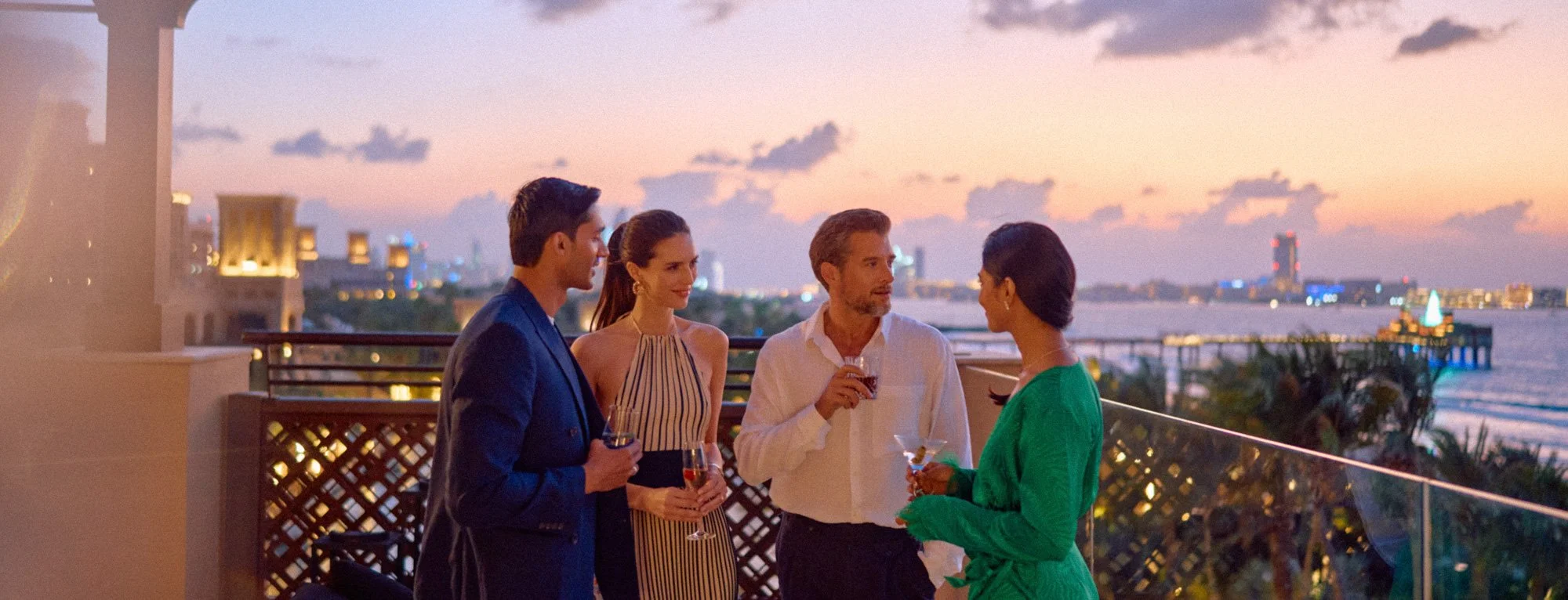 Four people enjoying drinks and conversation on a rooftop terrace during sunset, overlooking a city skyline with water in the background.