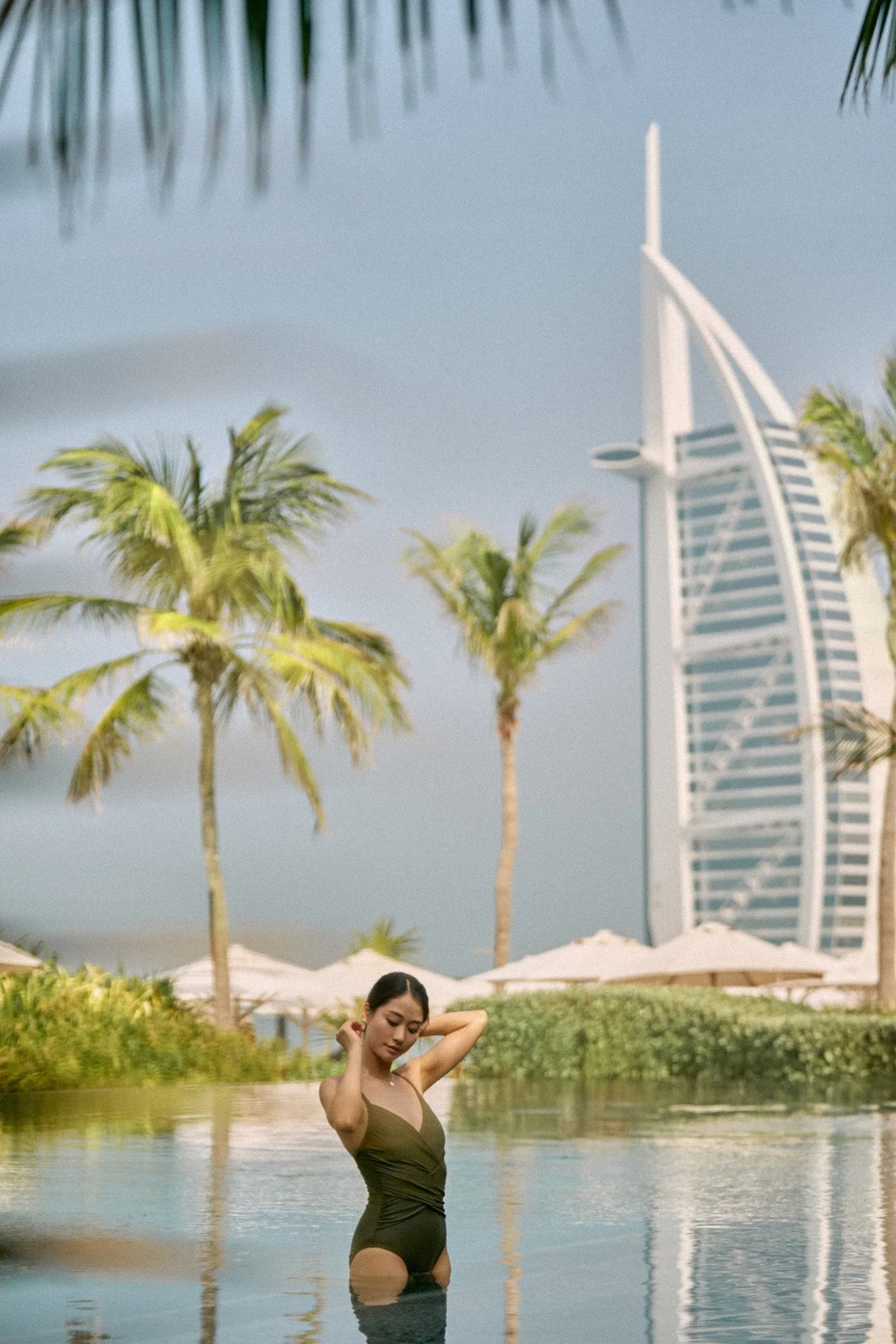 A woman in a swimsuit standing in a pool, with palm trees, umbrellas, and the Burj Al Arab hotel in Dubai visible in the background.
