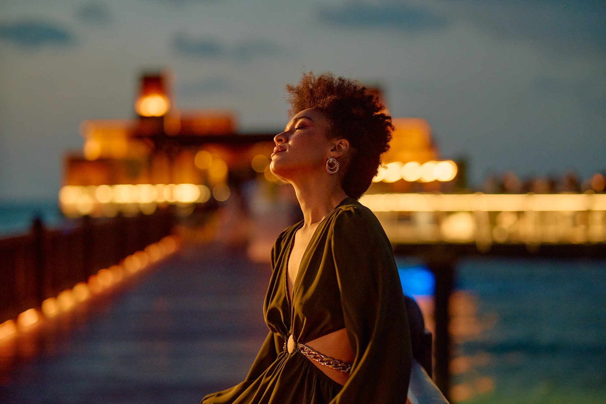Woman with closed eyes and joyful expression, standing on a pier at sunset with blurred lights and a building in the background.
