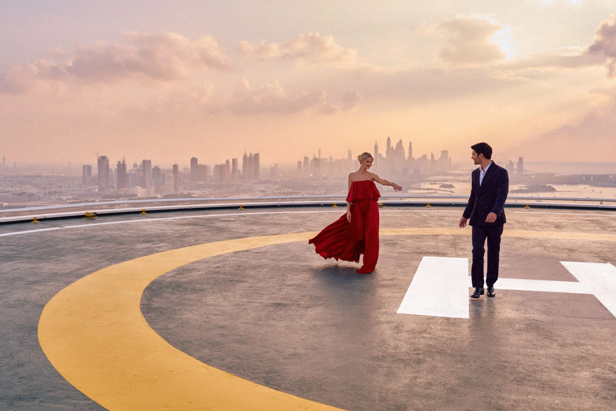 A woman in a red dress and a man in a suit dancing on the helipad of a skyscraper with a city skyline in the background during sunset.