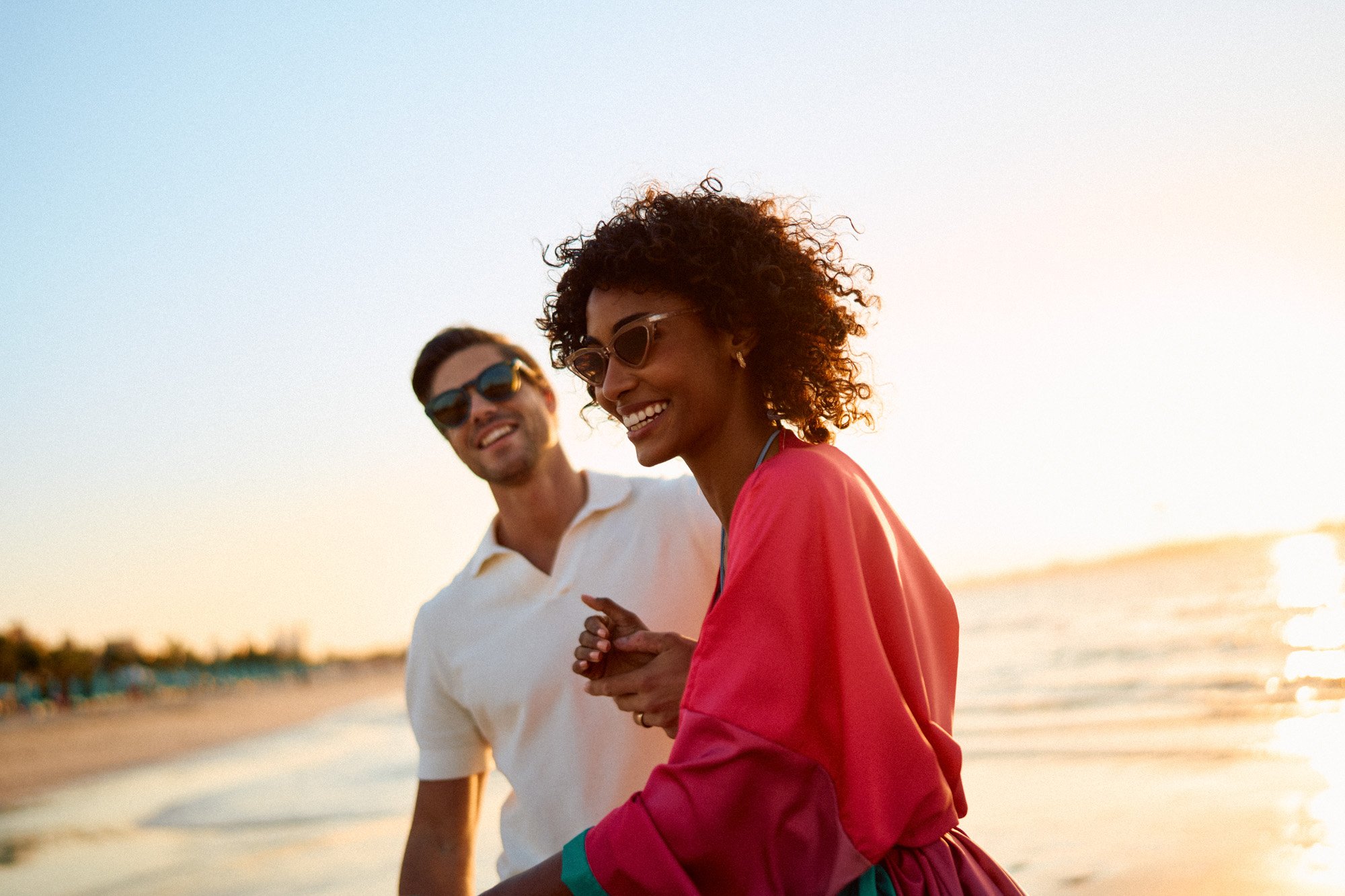 A man and woman walking on a beach at sunset, smiling and wearing sunglasses.
