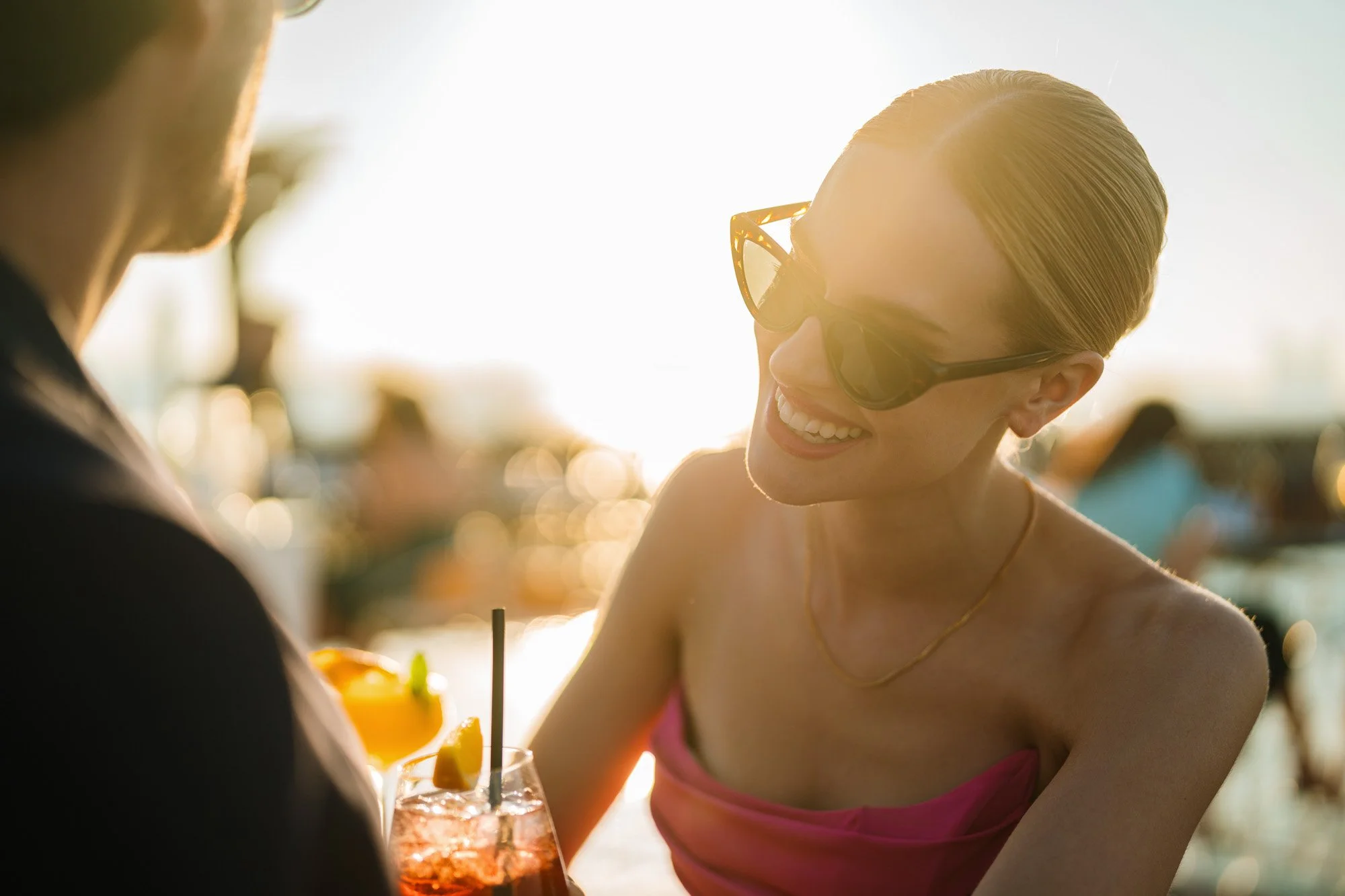 A woman in sunglasses smiling and talking to someone at an outdoor bar or restaurant during sunset, with a drink in front of her.