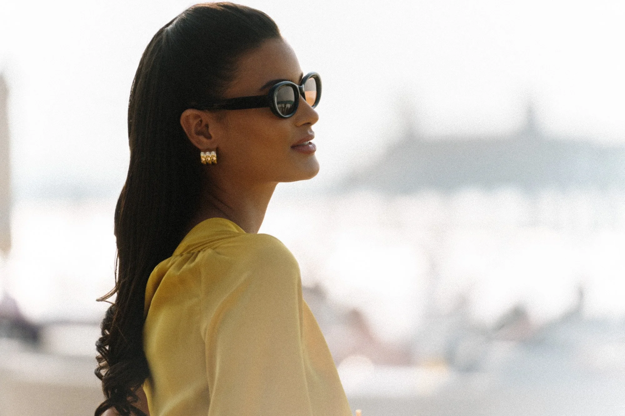A woman with long dark hair wearing black sunglasses, gold earrings, and a yellow top, looking to the side with a blurred background of water and a distant structure.