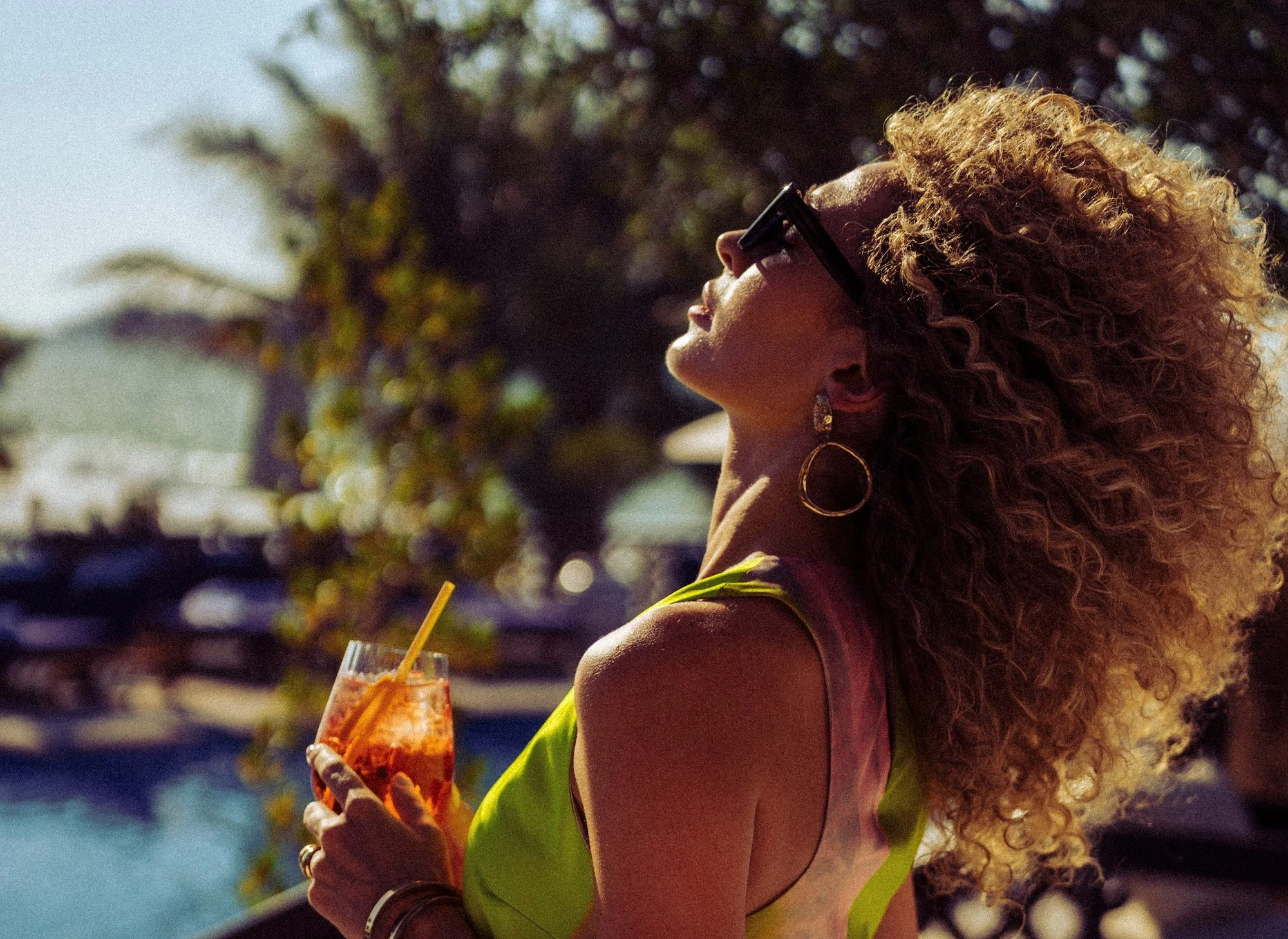 A woman with curly hair, wearing sunglasses and hoop earrings, holding a cocktail glass with an orange drink and a straw outdoors during daytime, with a blurred background of trees and water.