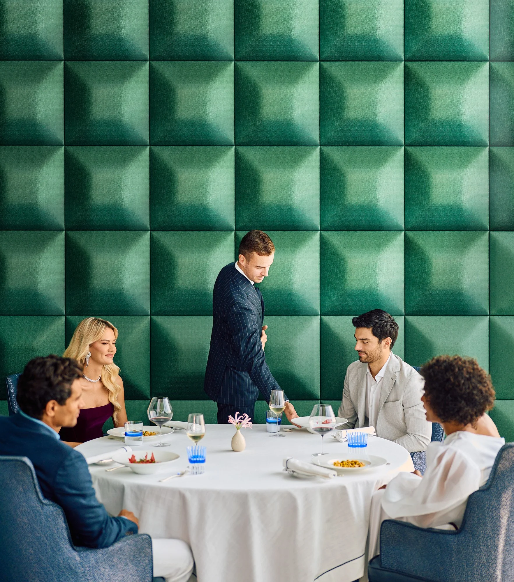 A man in a pinstripe suit shaking hands with a seated man at a round dining table set for a meal, surrounded by three other seated people in formal attire, in a restaurant with a green cushioned wall.