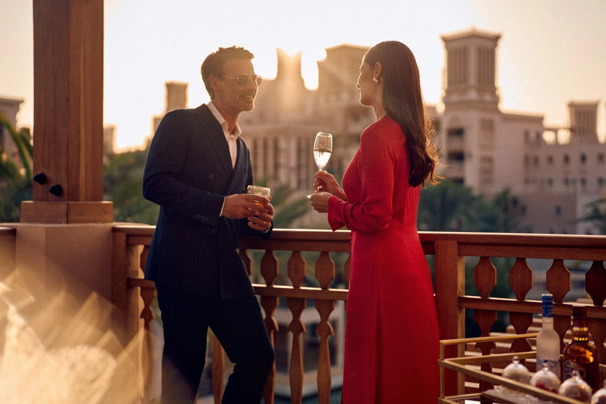 A man and woman talking on a balcony during sunset. The man is wearing a suit and sunglasses, holding a drink, and the woman is in a red dress, holding a glass of champagne. There are buildings in the background.