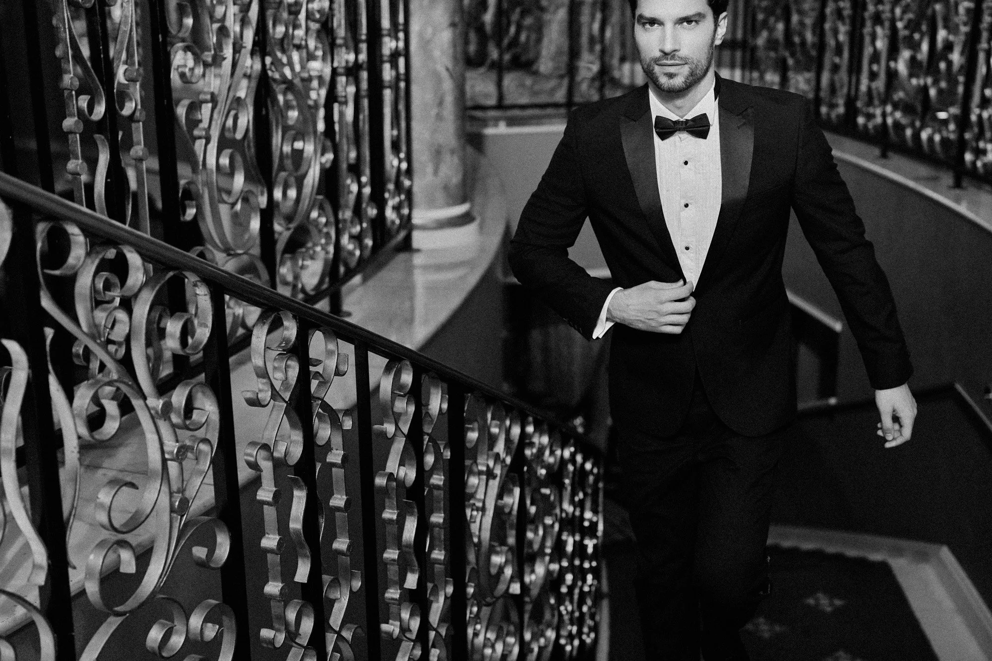 A man in a tuxedo with a bow tie walking up a staircase with ornate iron railings, in a formal setting.