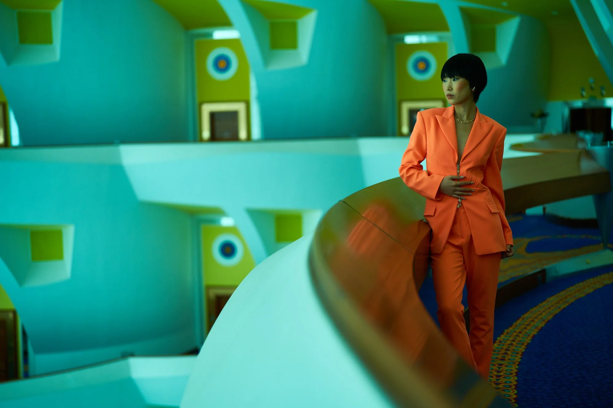 A woman in an orange suit standing in a colorful, retro-style room with curved architecture and patterned walls.