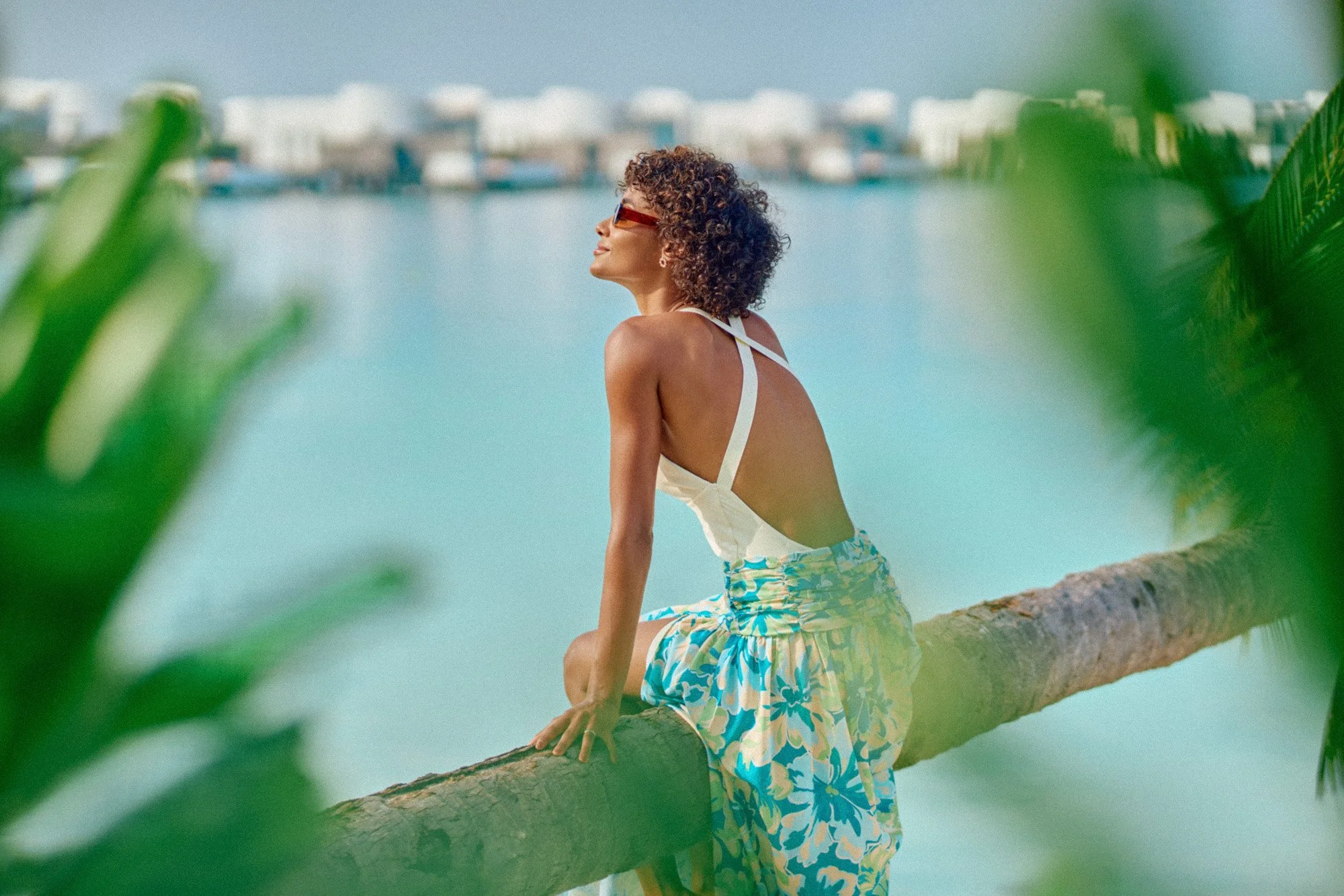 A woman with curly hair wearing sunglasses, a white backless top, and a colorful floral skirt, sitting on a tree branch near water, with a cityscape in the background.