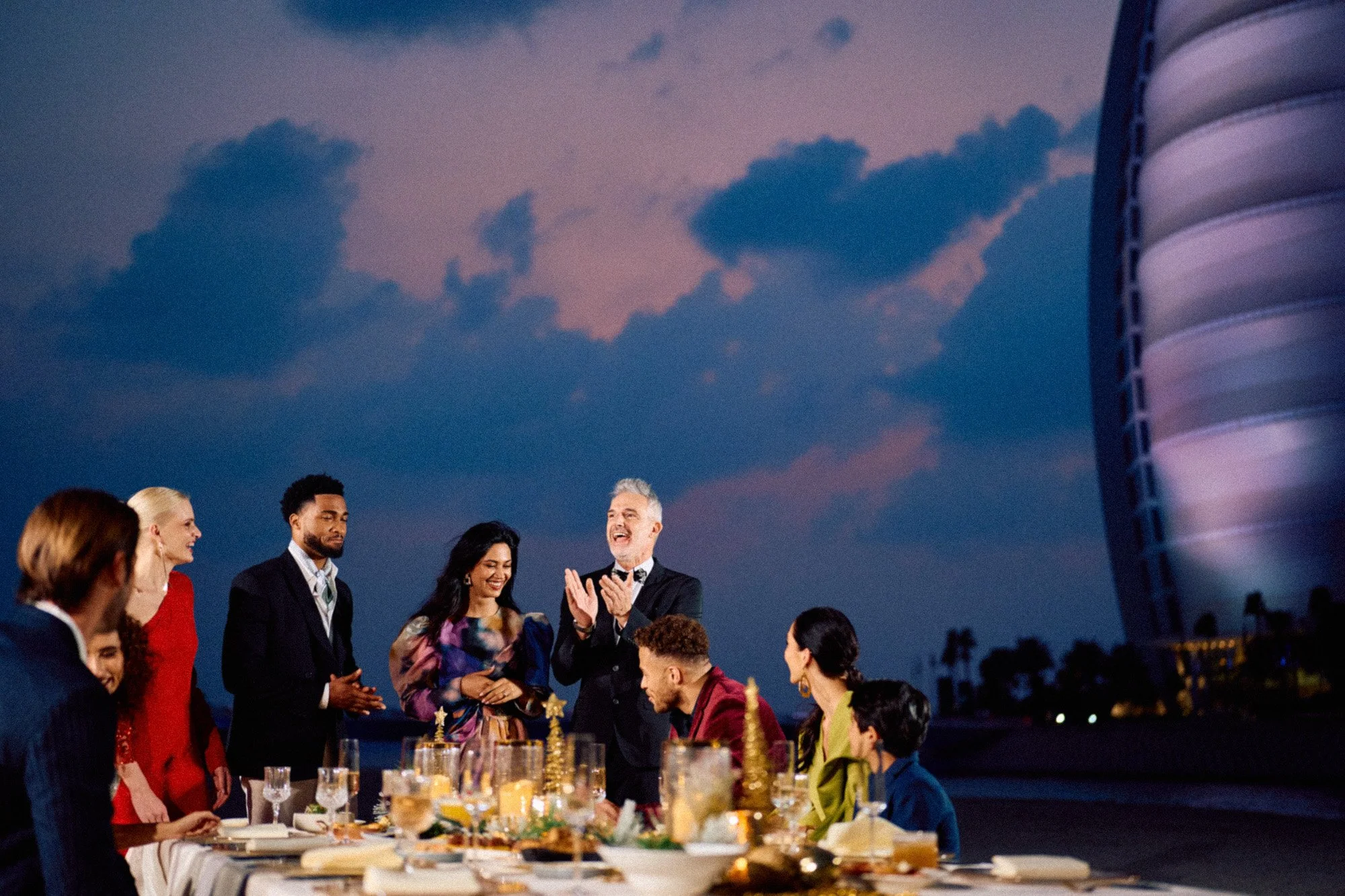 People gather around a table for an outdoor celebration during sunset, with a man in a suit clapping and smiling, and other diverse guests smiling and talking, with a modern building in the background.