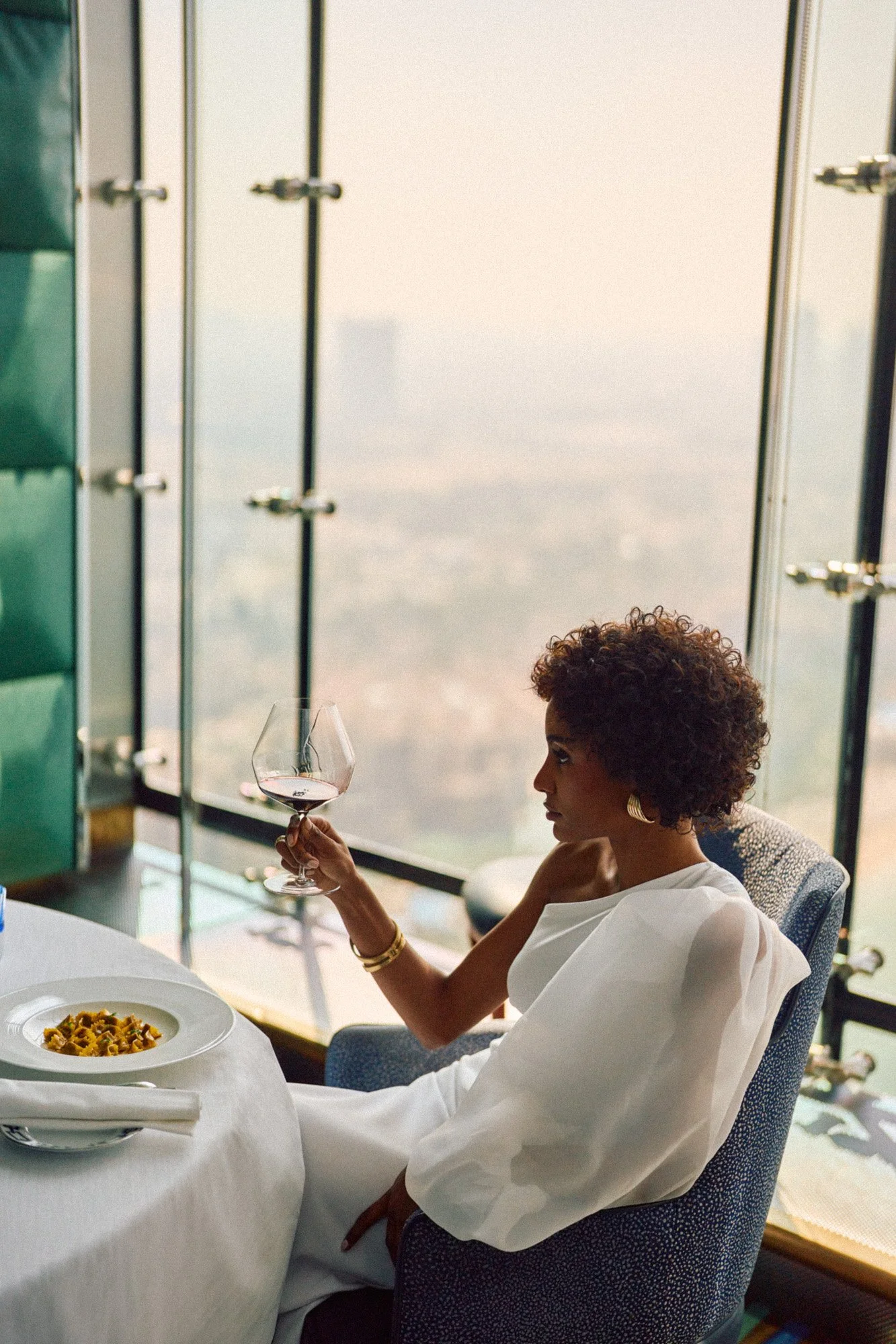 A woman with curly hair, wearing a white dress, sitting at a dining table with a plate of pasta, holding a glass of red wine, in a high-rise restaurant with large windows overlooking a city skyline.