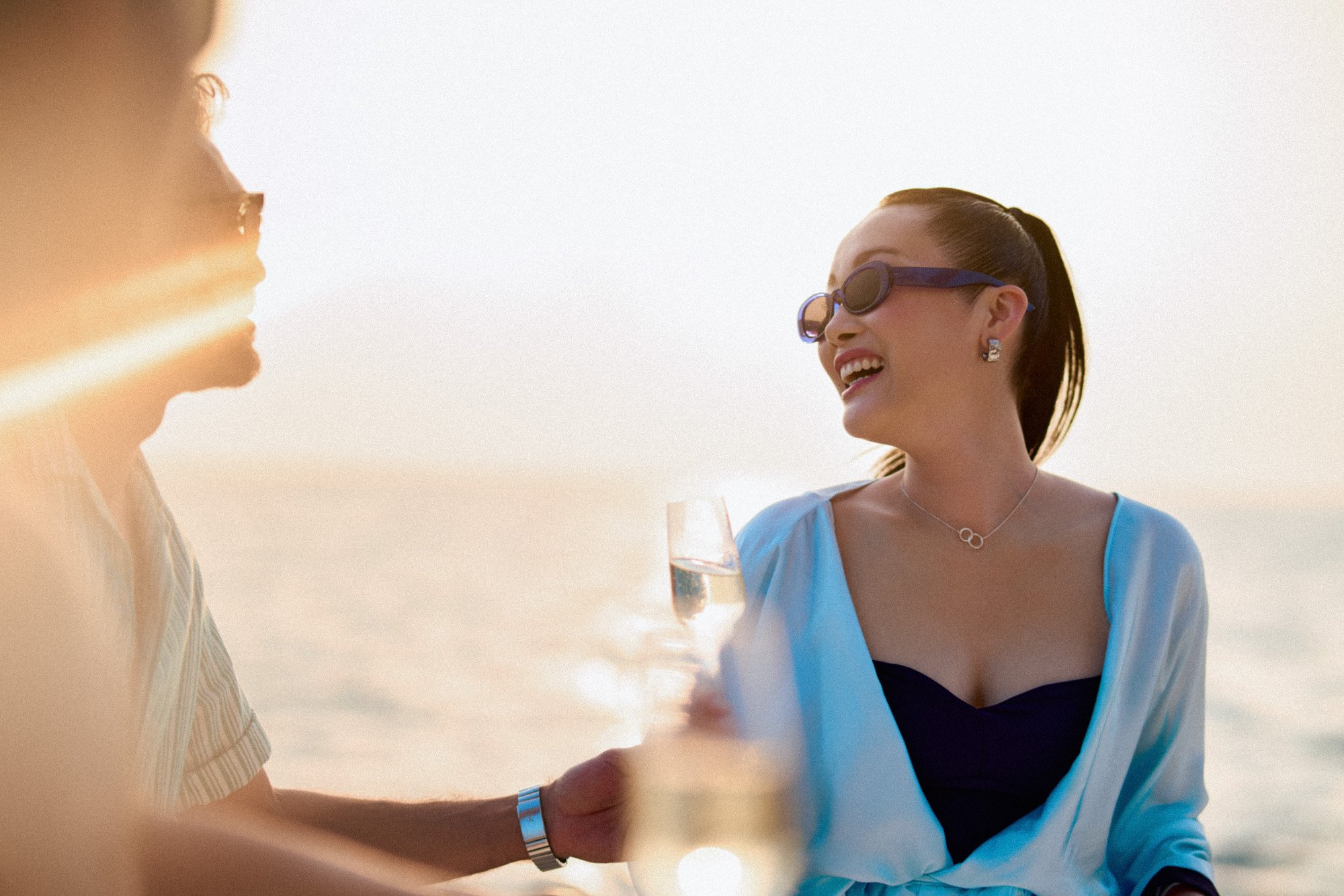 Two women laughing and chatting on a beach during sunset, holding champagne glasses.