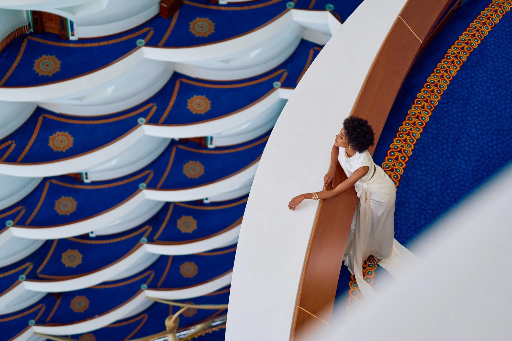 A woman in a white dress leaning on a wooden railing in an elegant theater or auditorium with blue and gold ornate ceiling decorations.