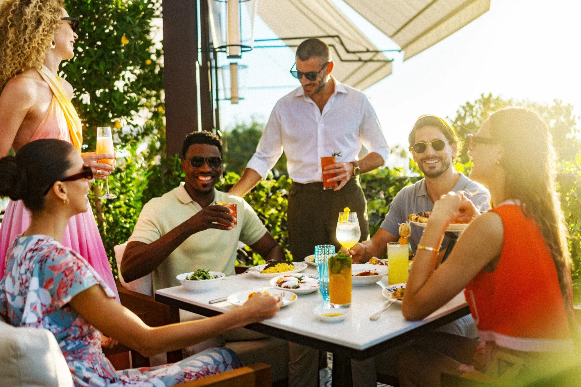Group of friends enjoying a summer outdoor meal at a restaurant, smiling and holding drinks, with food on the table.