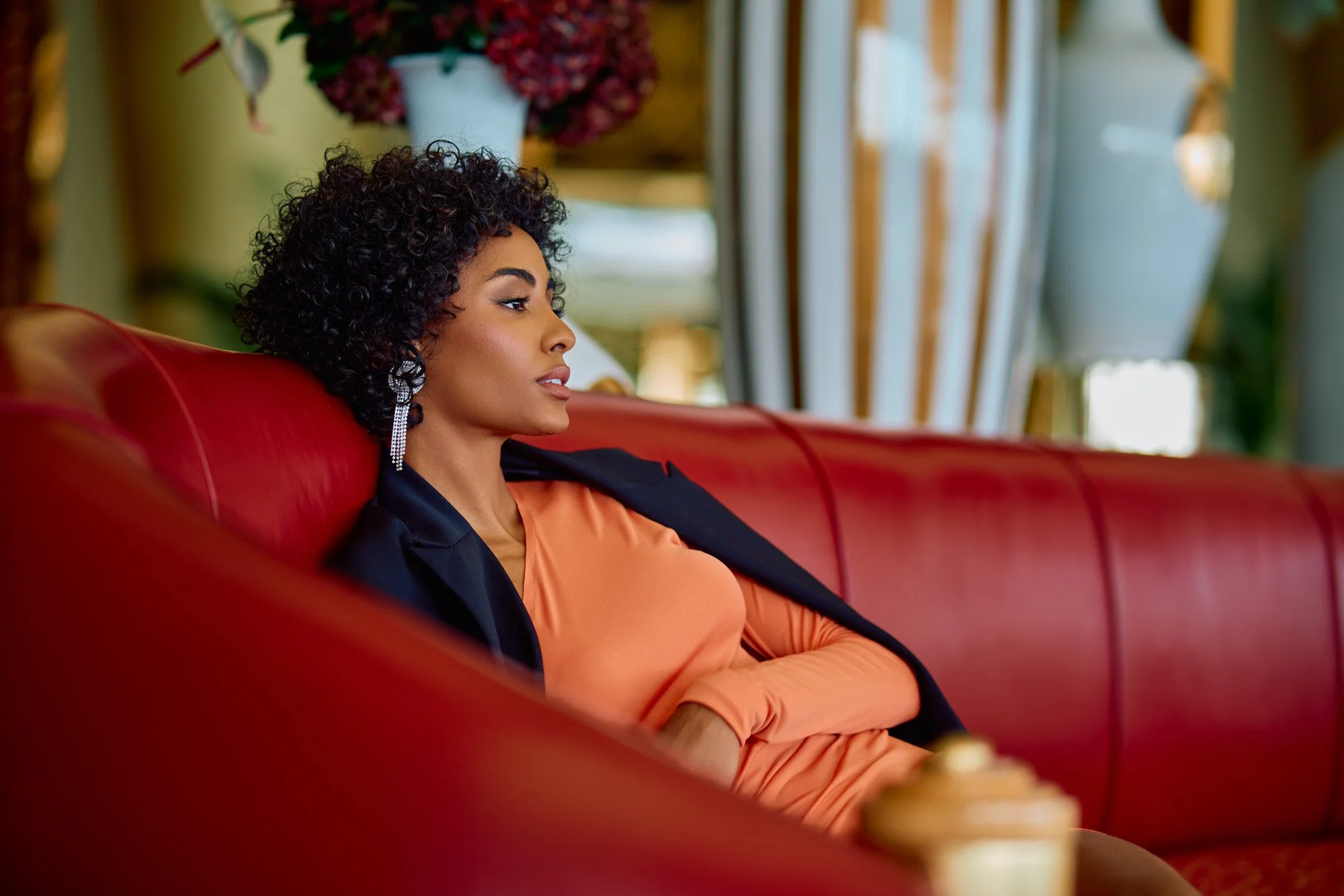 A woman with curly black hair, wearing earrings, a black blazer, and an orange dress, sitting on a red sofa in a luxurious setting with large flowers and decor in the background.