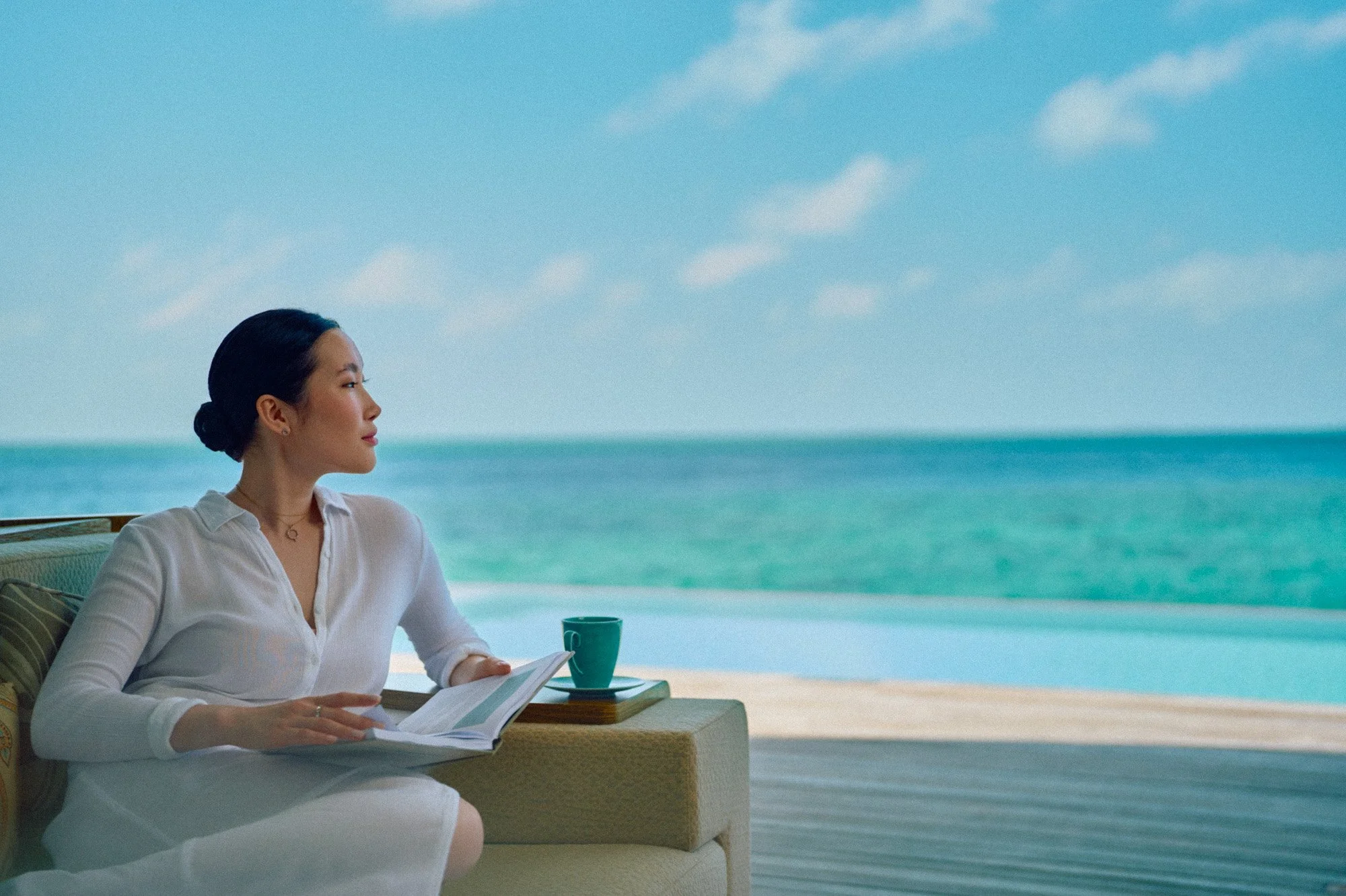 A woman sits on a sofa by the beach, holding a book, with a cup and a tablet nearby, overlooking the ocean under a blue sky with clouds.