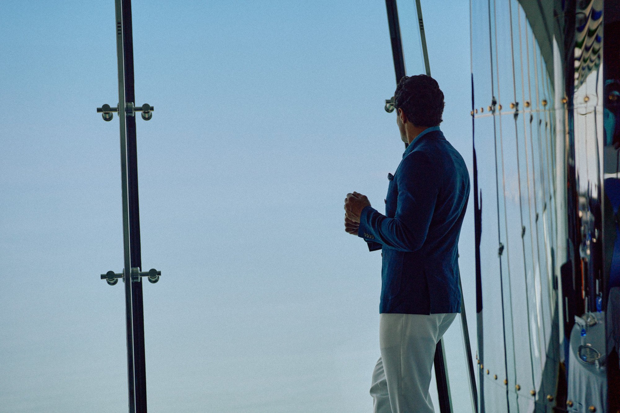 A man in a blue blazer and light-colored pants standing outside, looking into the distance during daytime.