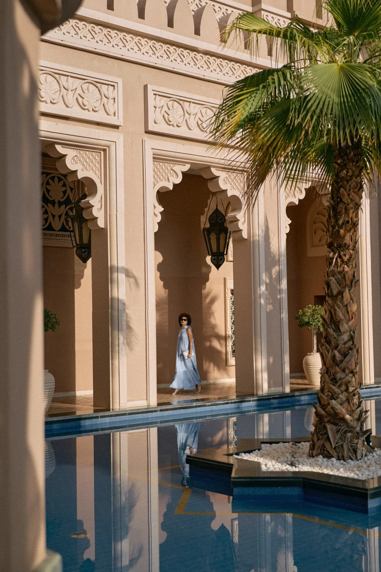 A woman in a light blue dress with sunglasses walking along a sunny courtyard with arches, ornate wall decorations, and palm trees reflected in a pool.