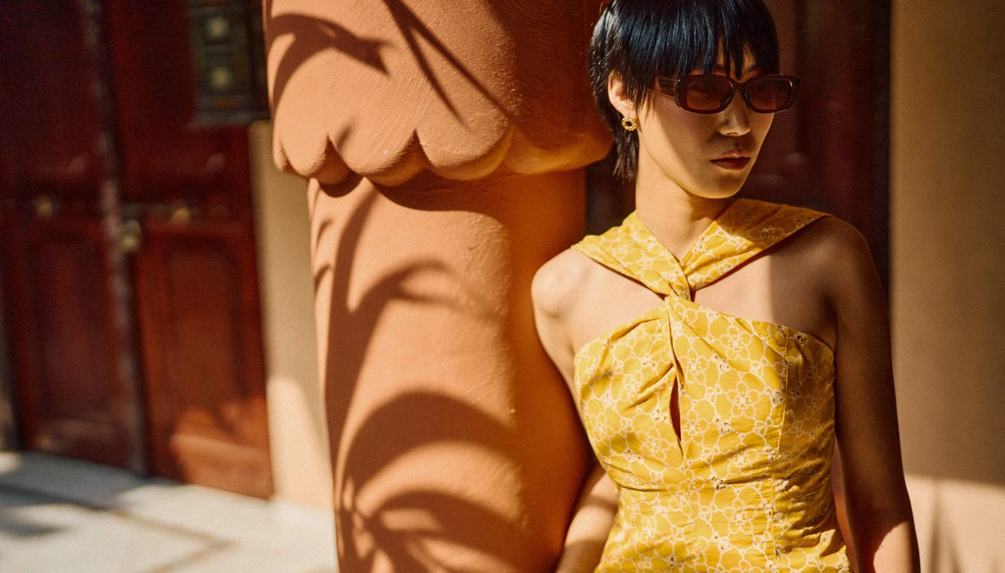 A woman wearing dark sunglasses and a yellow dress with a floral pattern, standing outdoors near a terracotta-colored column, with shadows cast by a plant's leaves on the column.