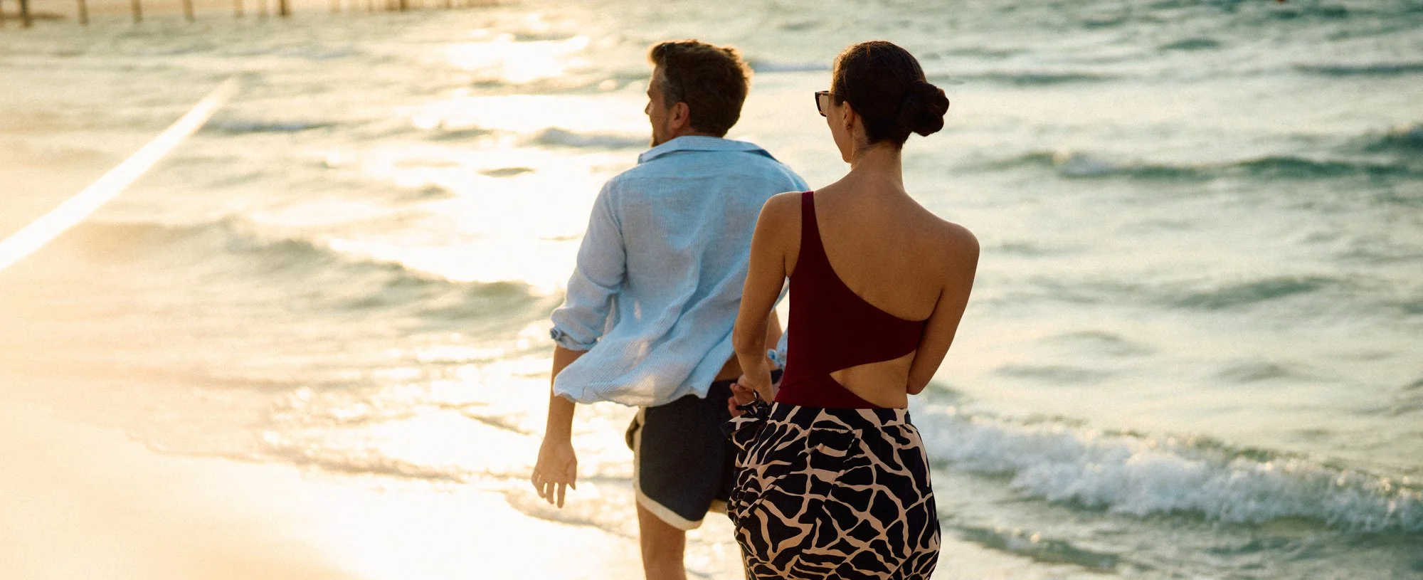 A man and woman walking along the beach at sunset, with the ocean waves in the background.