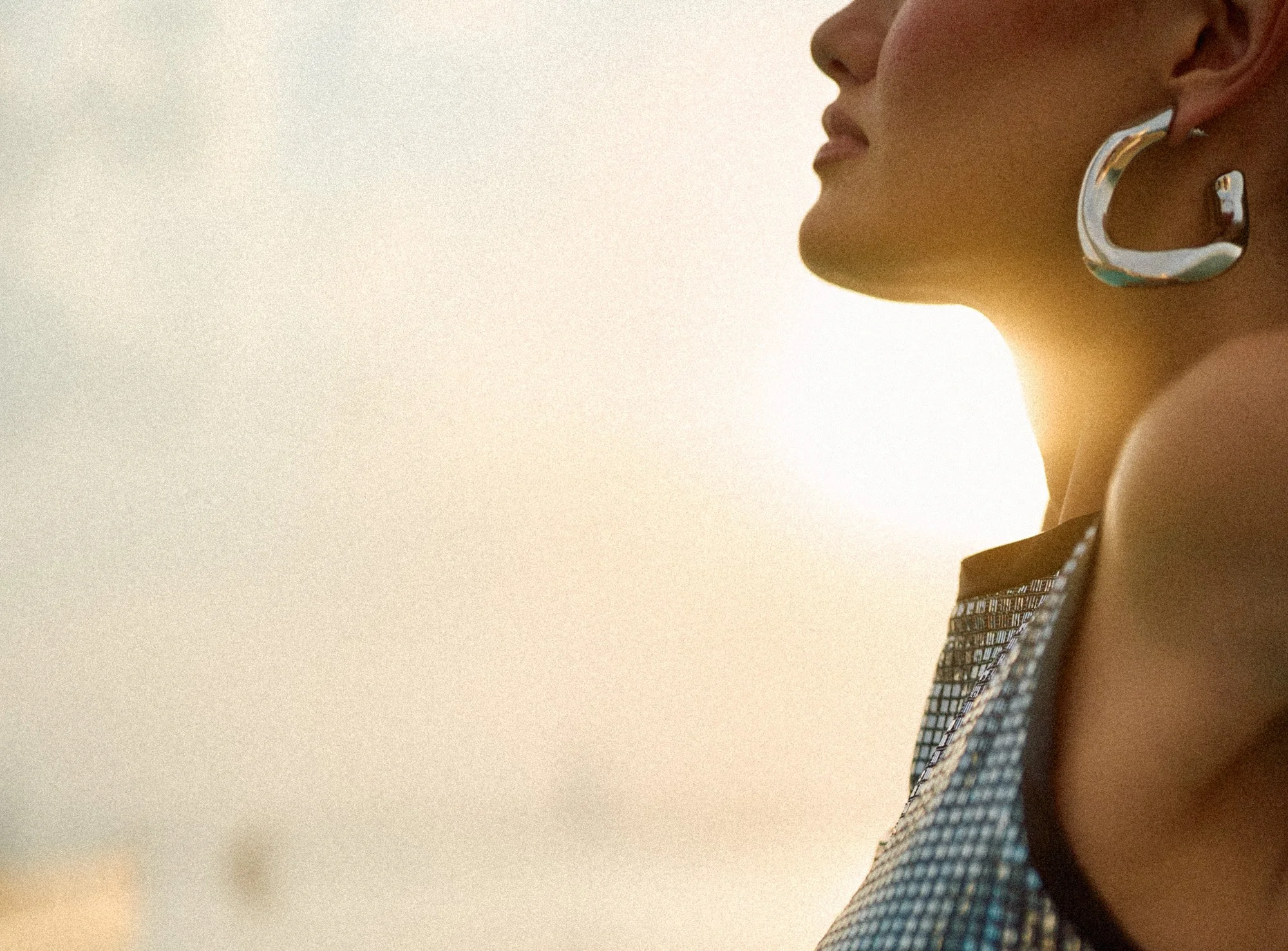 Close-up side profile of a woman wearing large silver hoop earrings and a patterned sleeveless top, with warm sunlight illuminating her face and shoulder.