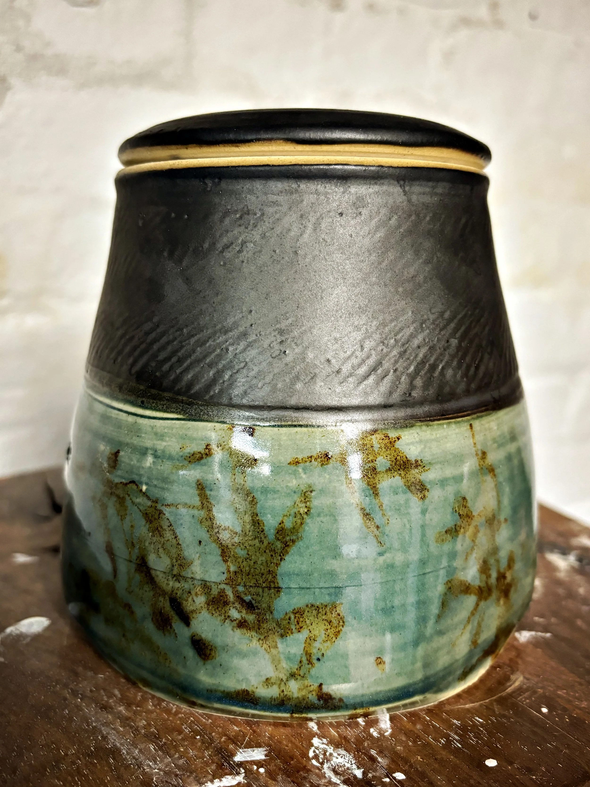 A ceramic vase with a matte black upper section and a greenish blue lower section featuring a textured, crackled glaze and a colored, abstract pattern.