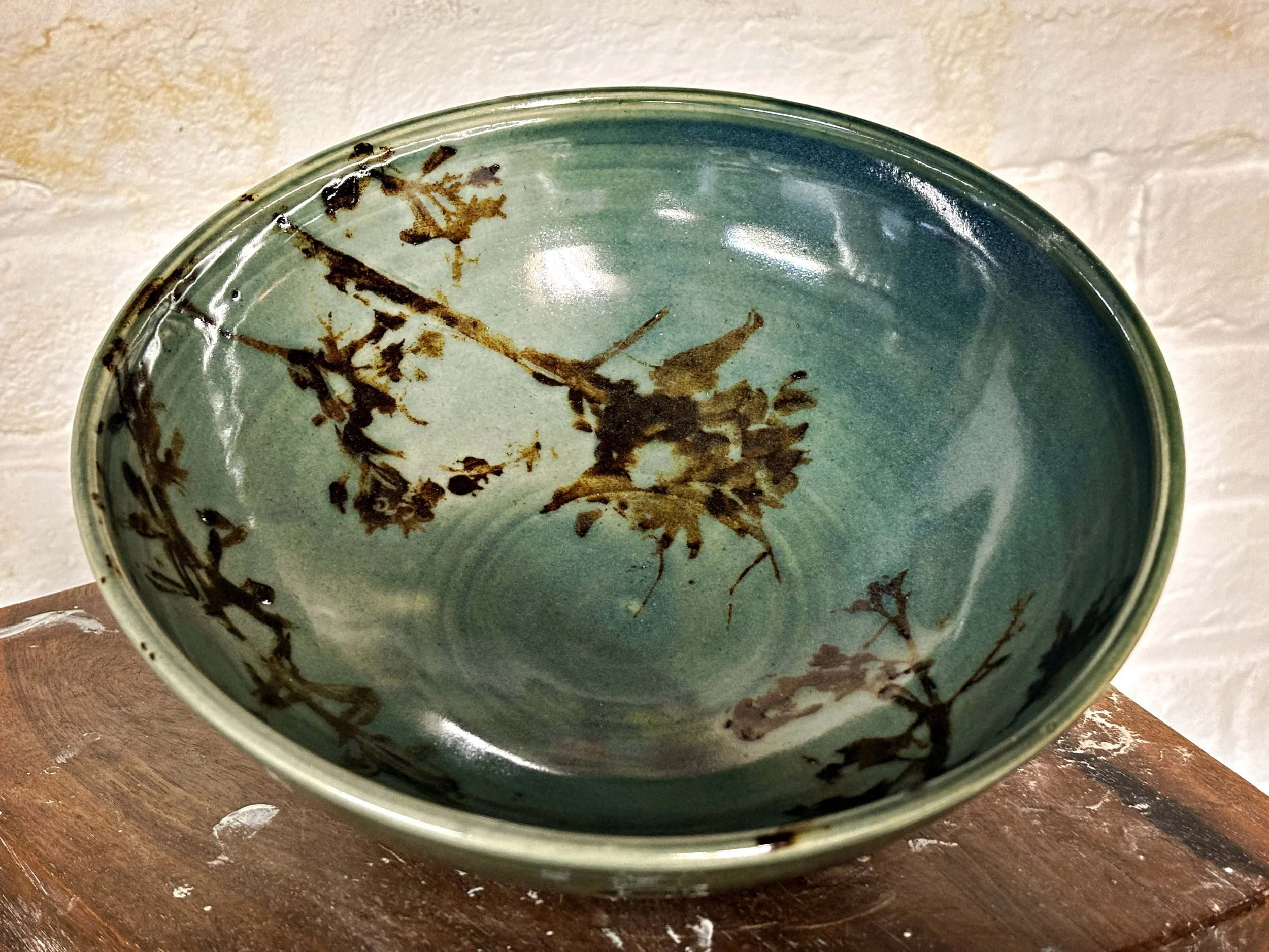 A ceramic bowl with green glaze and brown decorative plant patterns, placed on a wooden surface against a cream-colored wall.