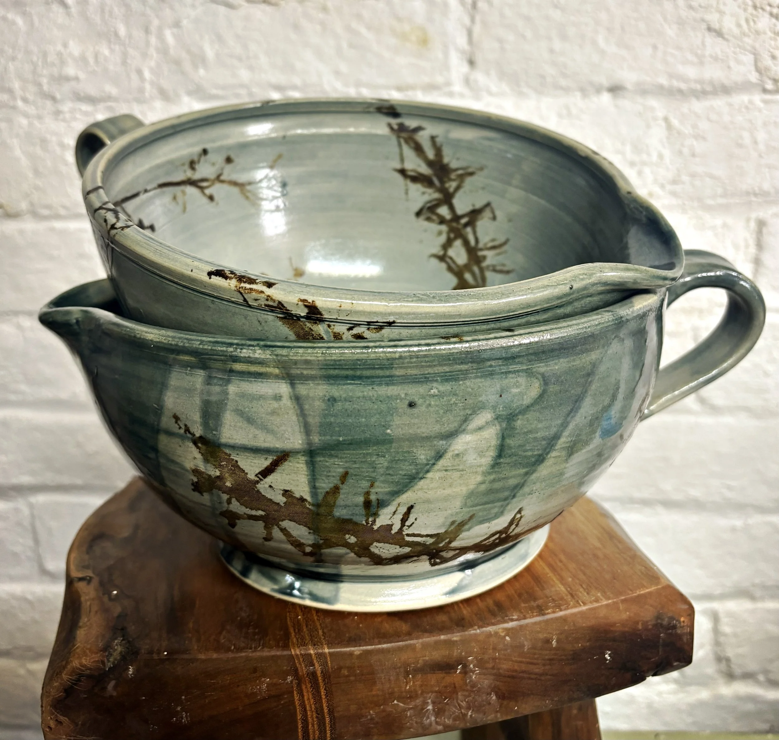 Two stacked ceramic batter bowls with hjandles and with a greenish glaze and brown decorative patterns, placed on a wooden surface against a white brick wall.