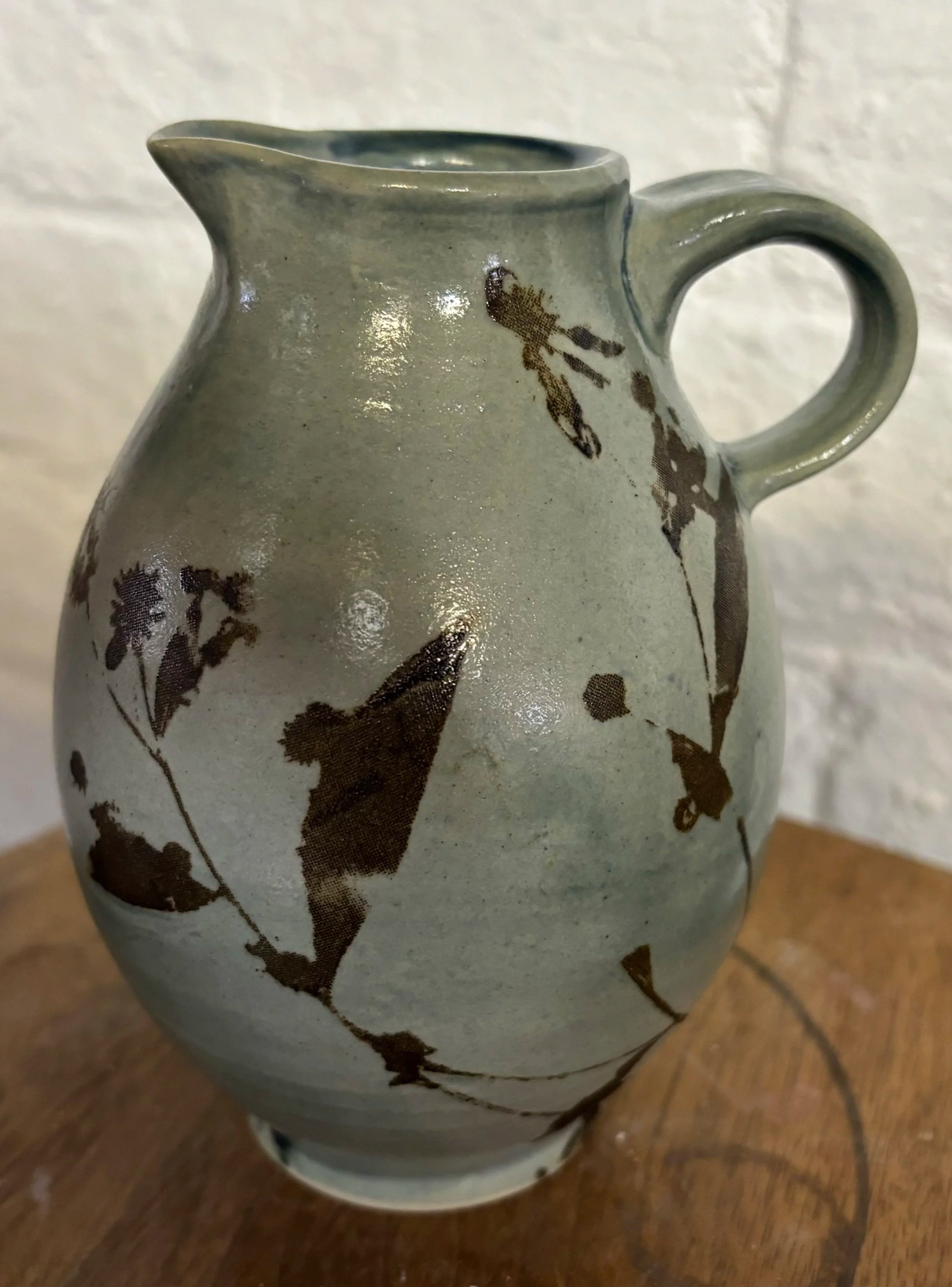 A ceramic vase with a handle, decorated with a dark floral pattern, placed on a wooden surface against a white textured wall.