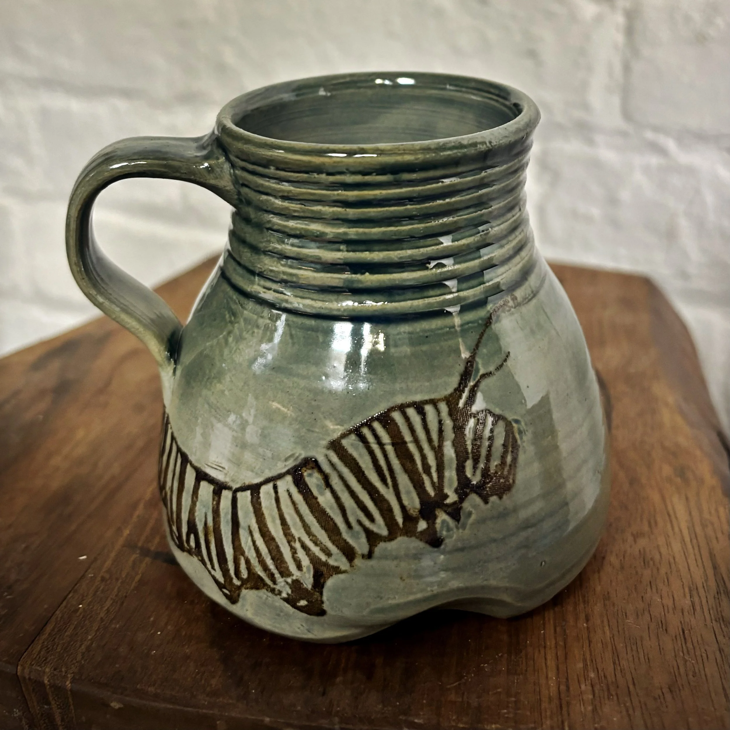 A handcrafted ceramic mug with green glaze and a brown insect design, featuring a handle and series of horizontal ridges near the top, placed on a wooden surface.