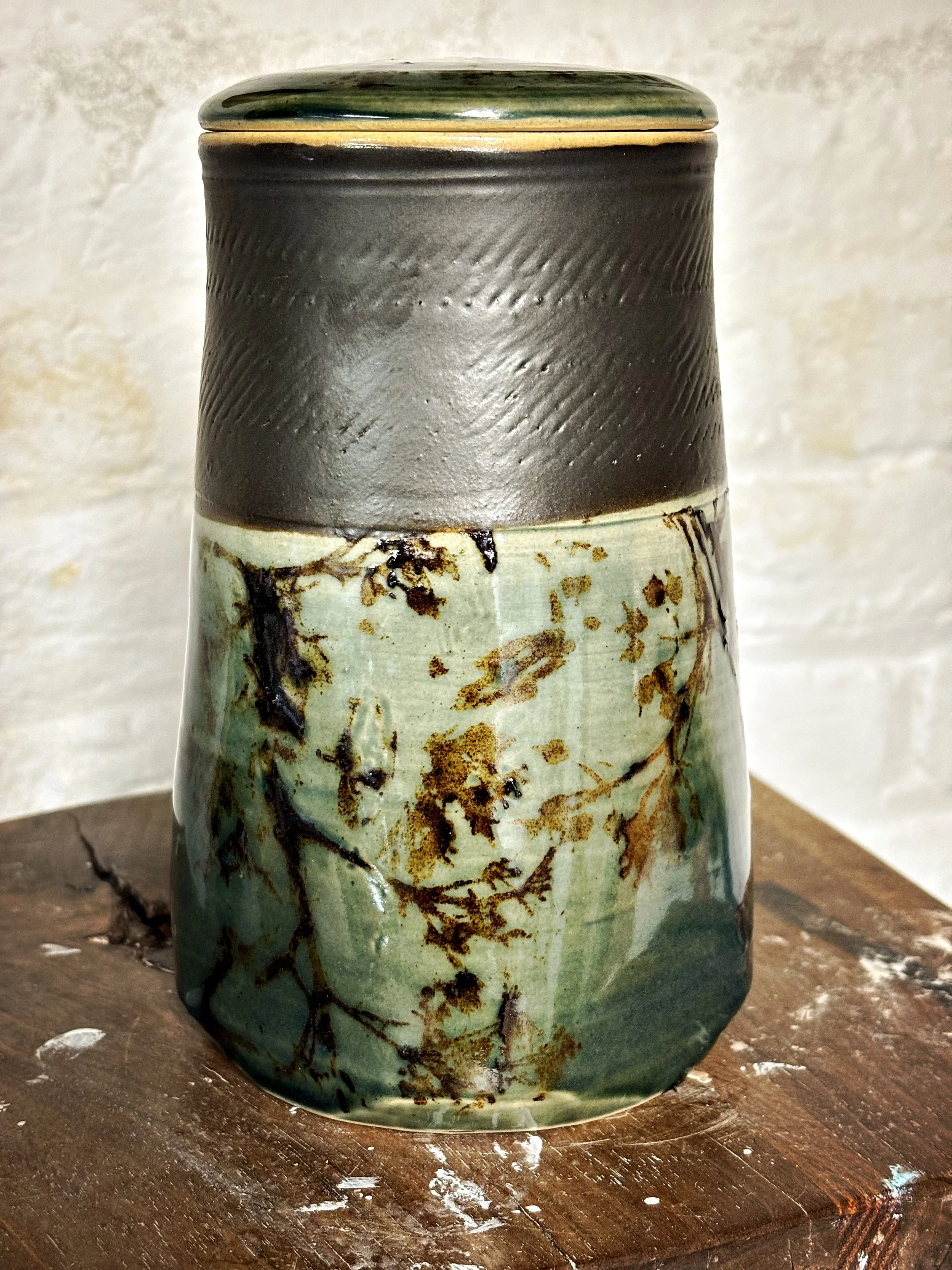 A handmade ceramic jar lidded with a dark upper section and a glazed lower section featuring a natural, abstract pattern in earthy tones, placed on a wooden surface.