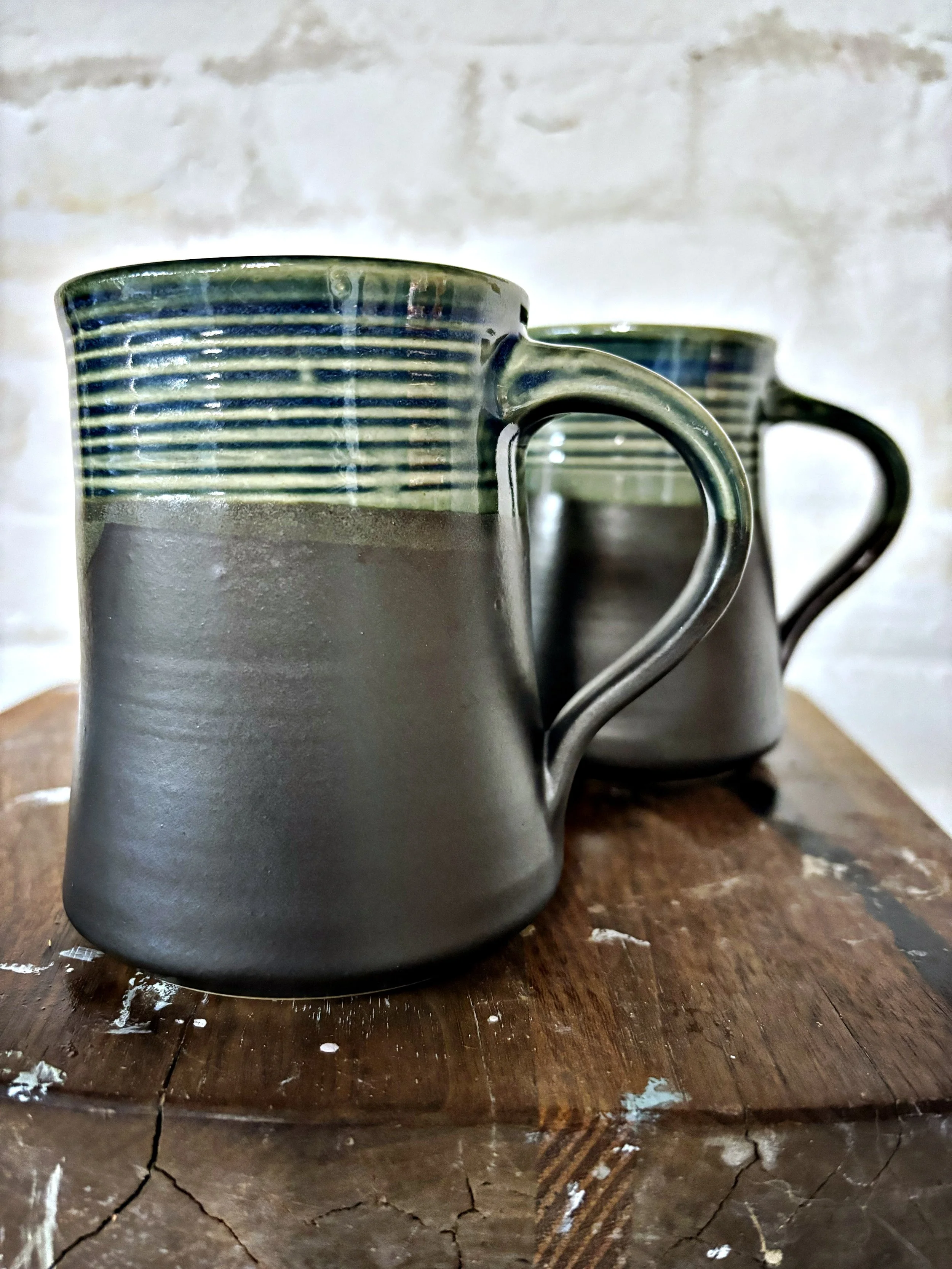 Two ceramic mugs with matyte black grey bases and green and blue striped tops, placed on a rustic wooden surface with a cracked finish, against a light-colored brick wall background.
