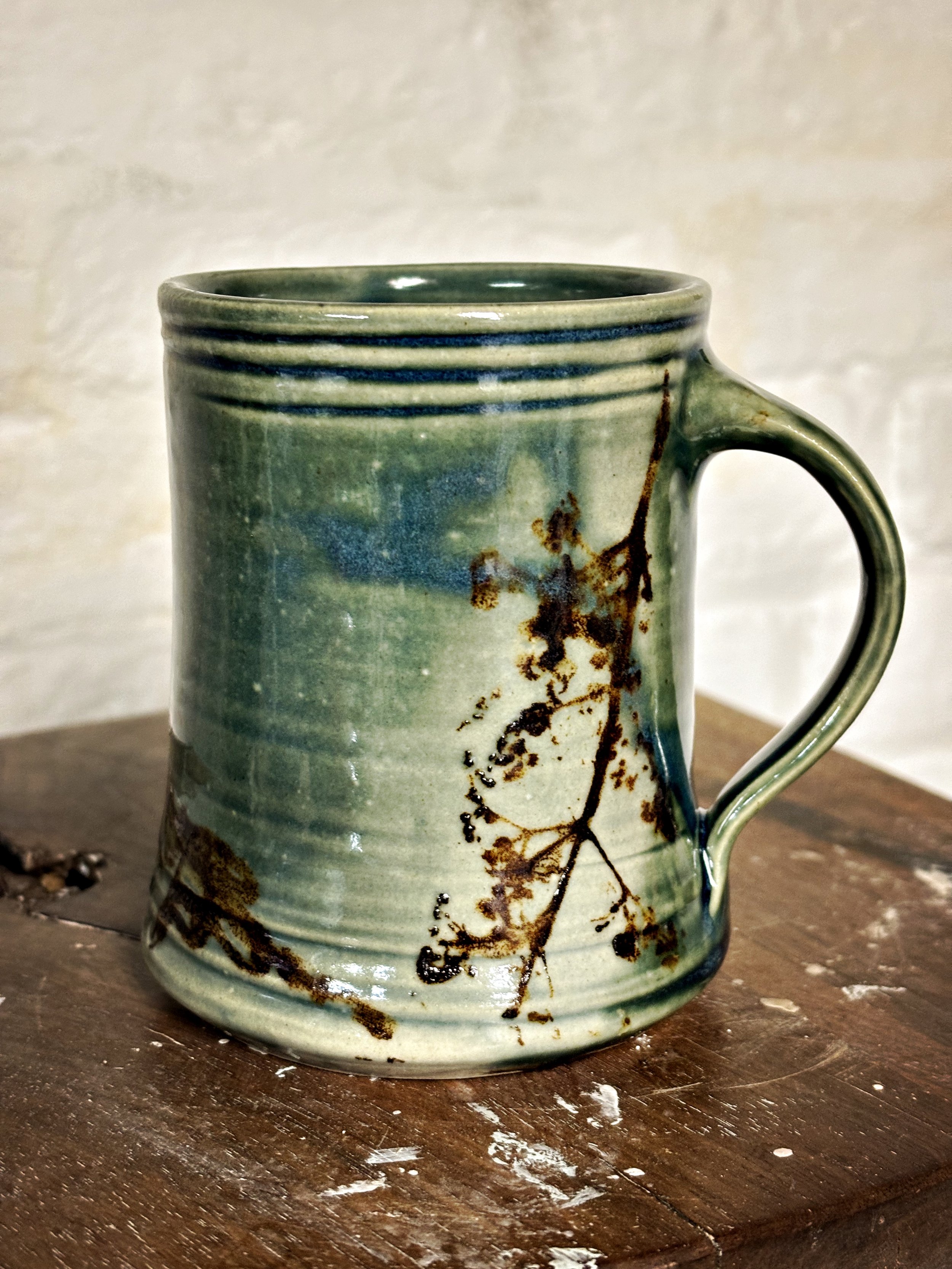 A green ceramic mug with brown leaf and branch design, placed on a wooden surface.