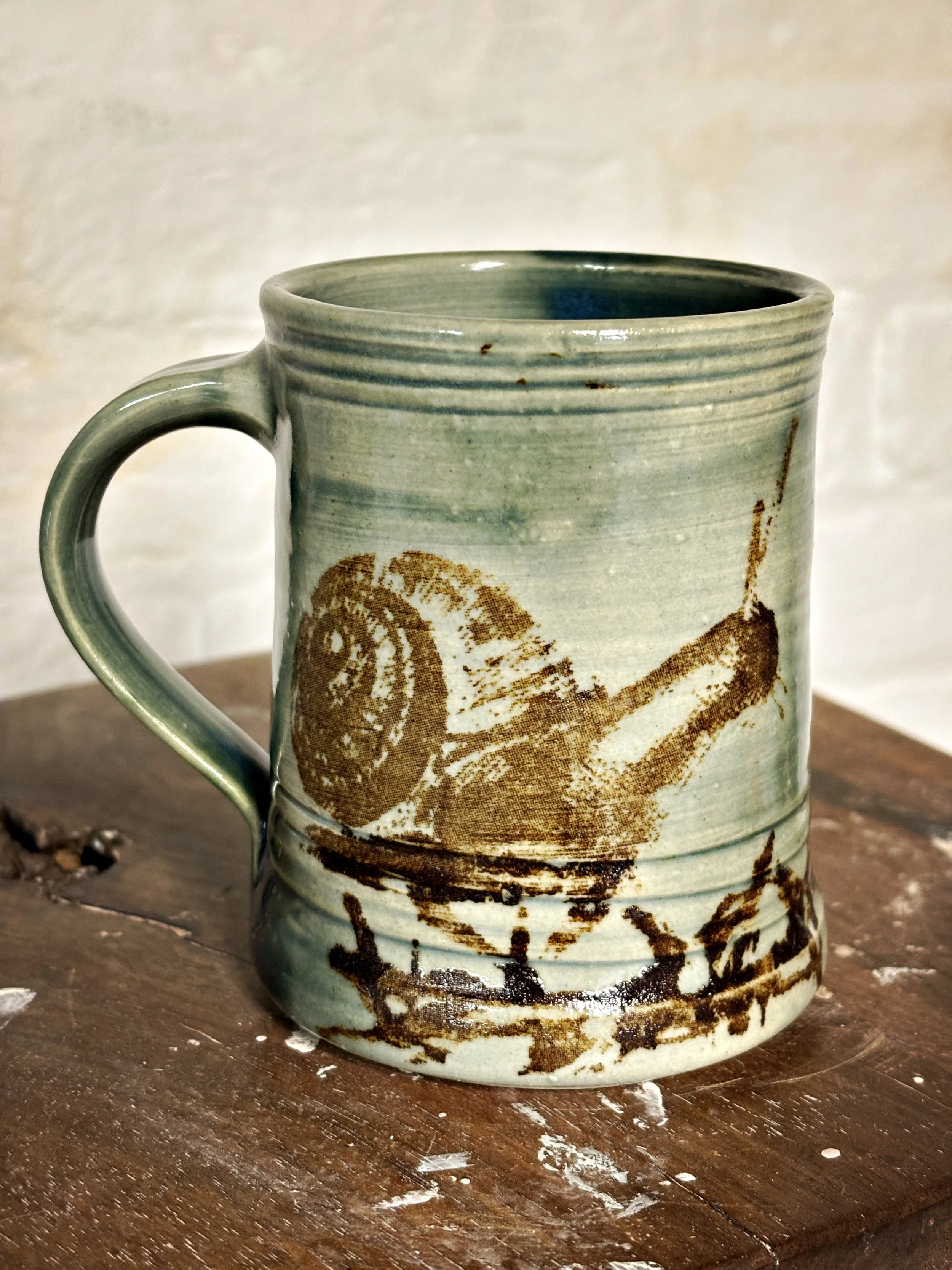 A ceramic mug with a greenish glaze and a brown design depicting a snail, placed on a wooden surface.