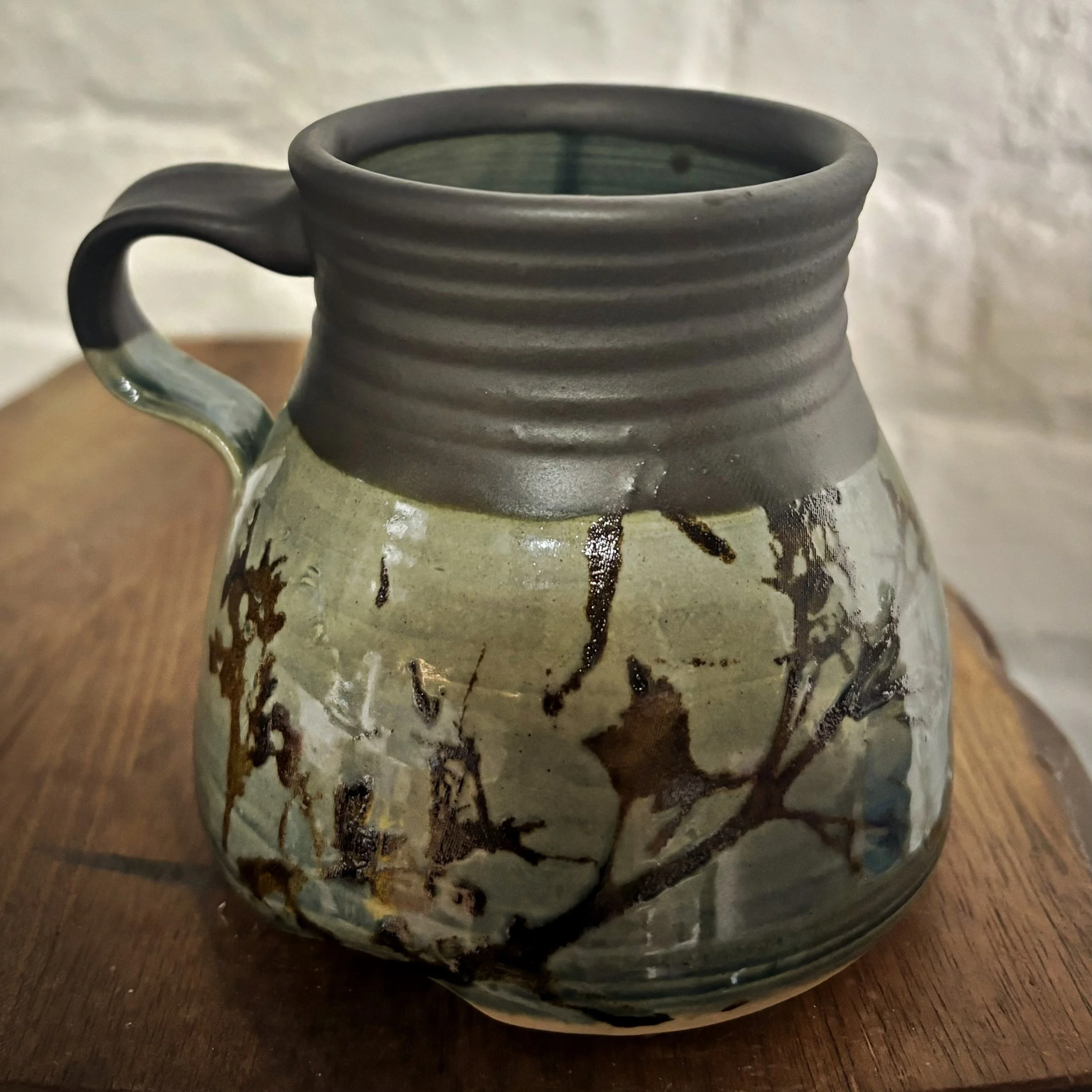 A ceramic mug with a matte black, ribbed upper section and a glazed lower section featuring a branch with leaves and flowers design in earthy tones, placed on a wooden surface.