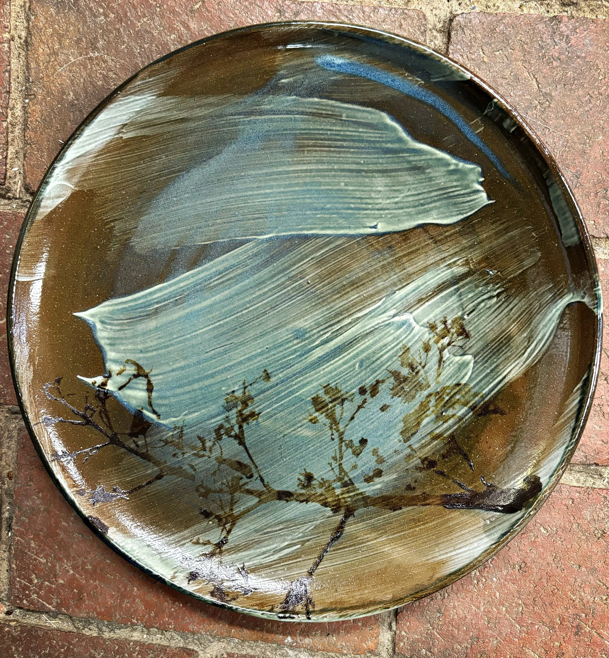 A round ceramic plate with a painted design, featuring abstract brushstrokes in shades of blue, green, and tan, and a silhouette of a branch with small leaves in darker brown, placed on a brick surface.