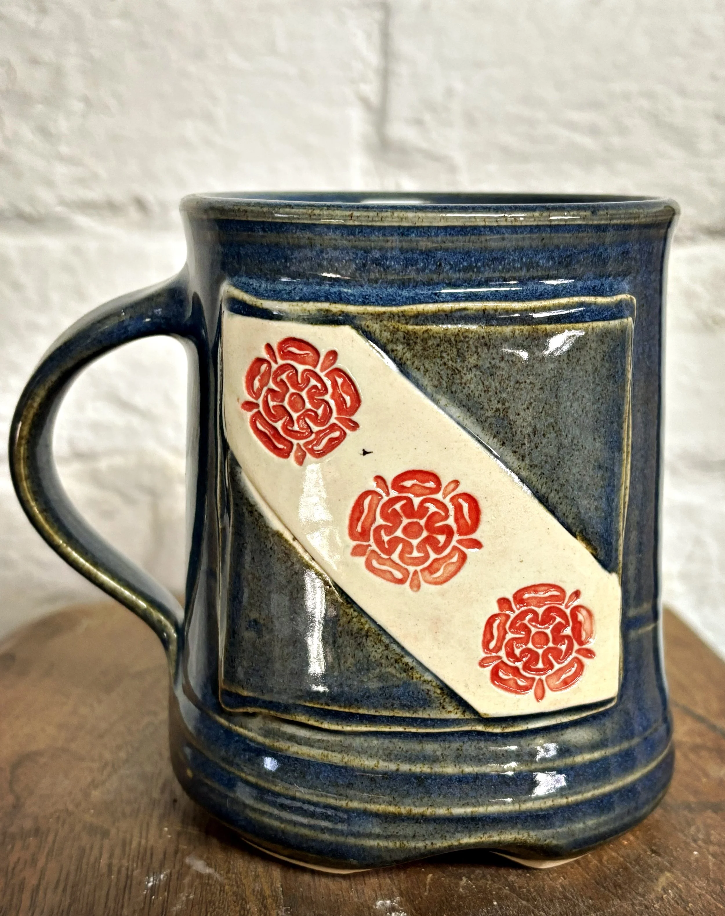 Ceramic mug with a blue, gray, and white geometric pattern, featuring Crozet Virginia flag with three red decorative floral motifs on a white diagonal stripe.