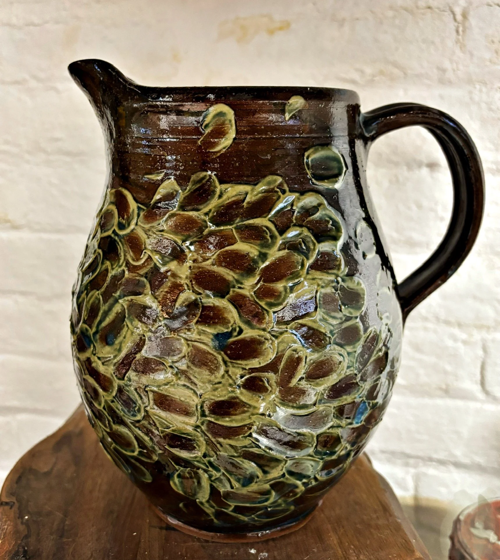 Brown ceramic pitcher with a textured surface resembling sunflower seeds, placed on a wooden surface against a light-colored brick wall.