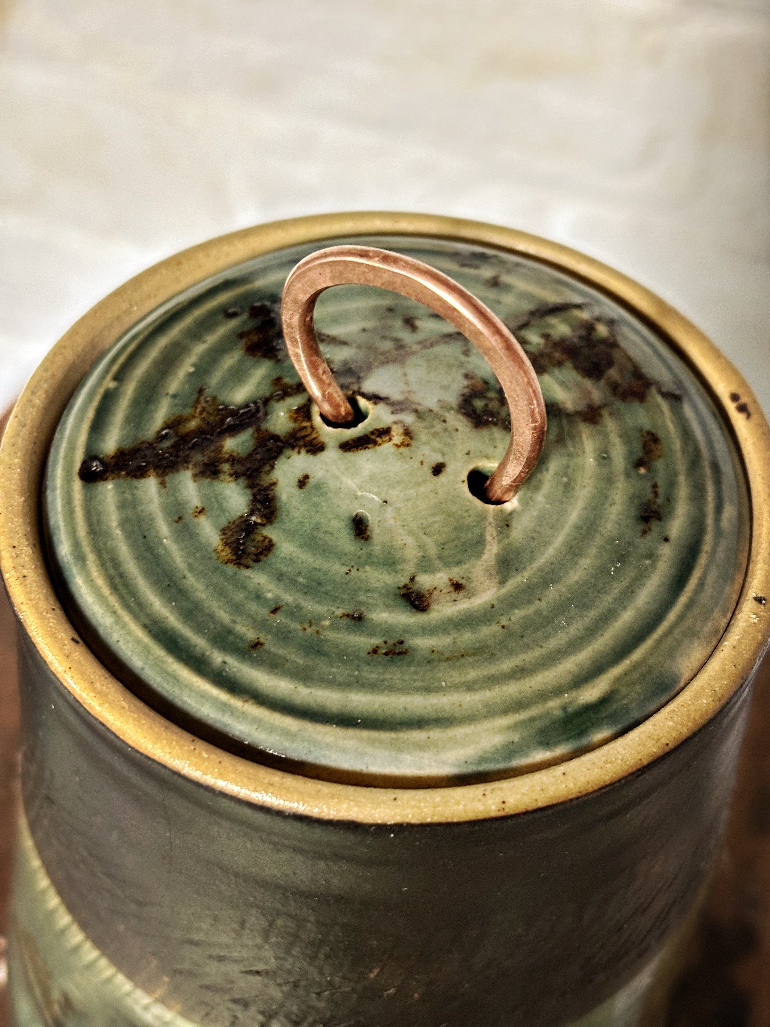 Close-up of a ceramic container with a copper lid pull handle on a leafy green and black stained ceramic lid.