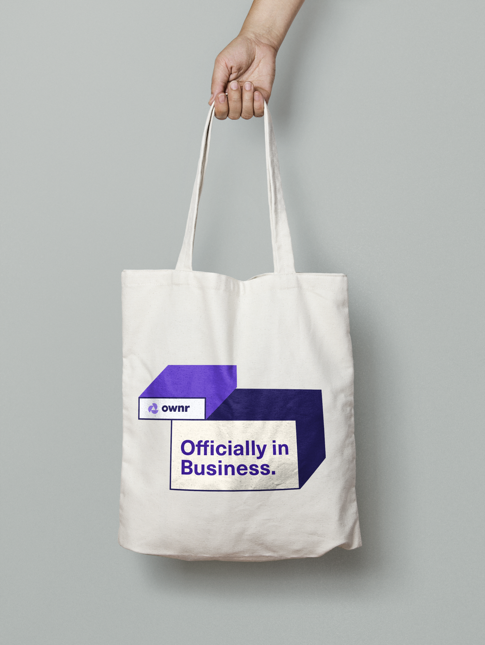 Hand holding a white tote bag with the text "Officially in Business" and the logo "ownr" on a plain background.