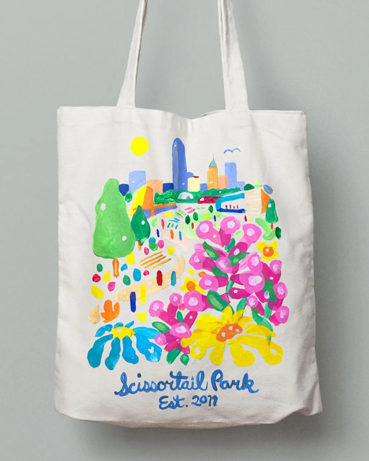 Colorful tote bag with a hand-painted illustration of a city park with flowers, trees, and a skyline, and the text "Scissortail Park Est. 2019".