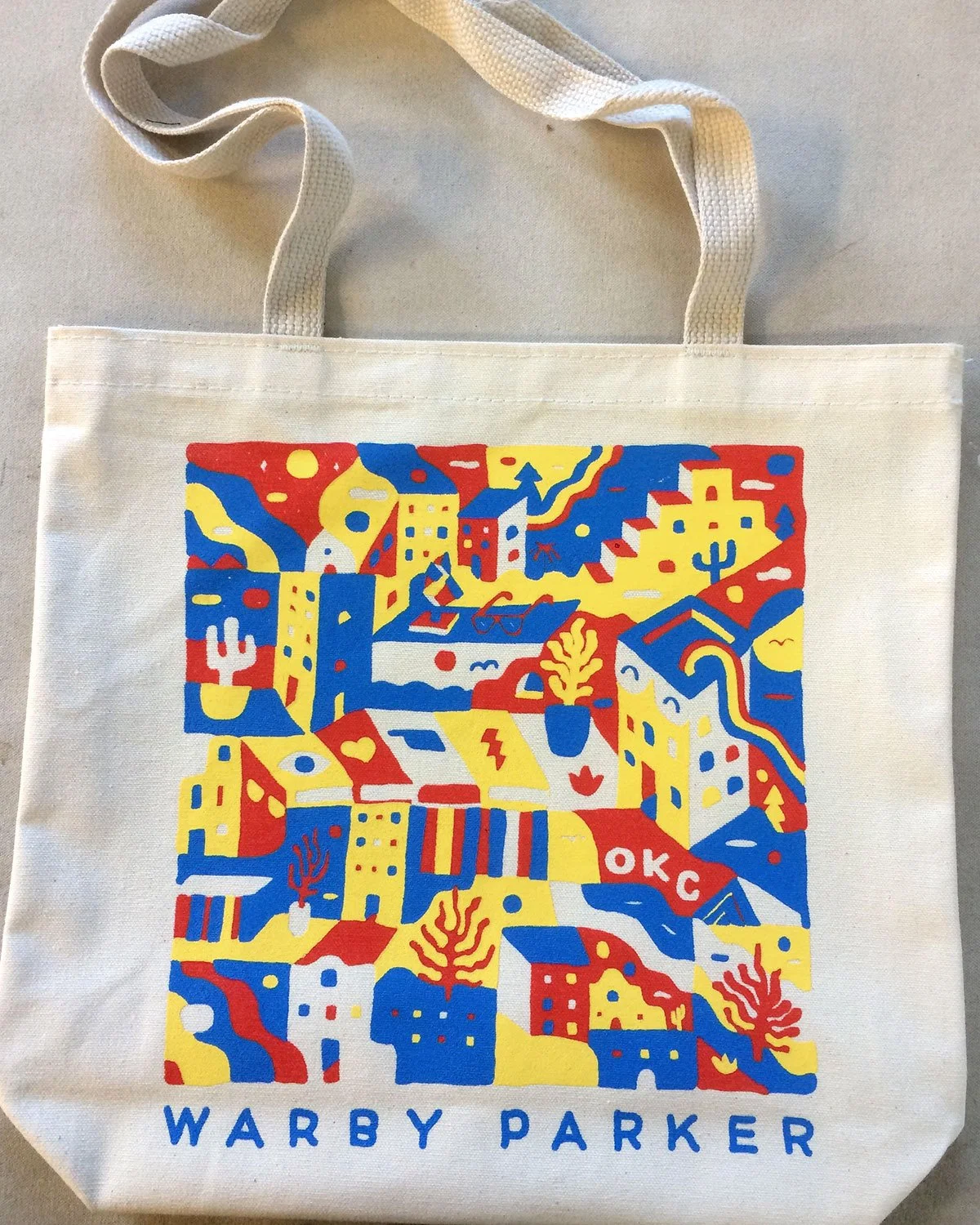 A white tote bag featuring a colorful, stylized illustration of a cityscape with buildings, mountains, and cacti in red, blue, yellow, and white. The text "WARBY PARKER" is printed at the bottom of the bag.